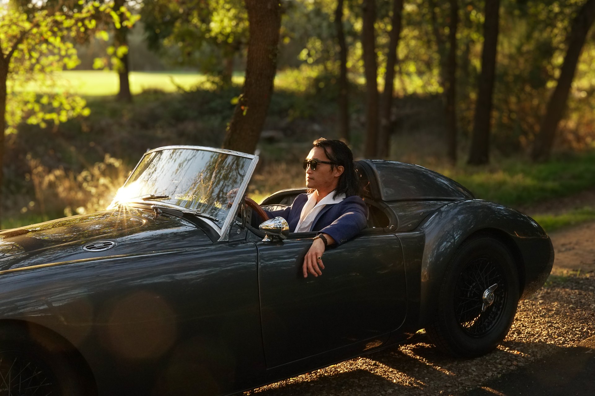 A photo of a man sitting in the drivers seat of a 1958 MG MGA in a park setting during golden hour