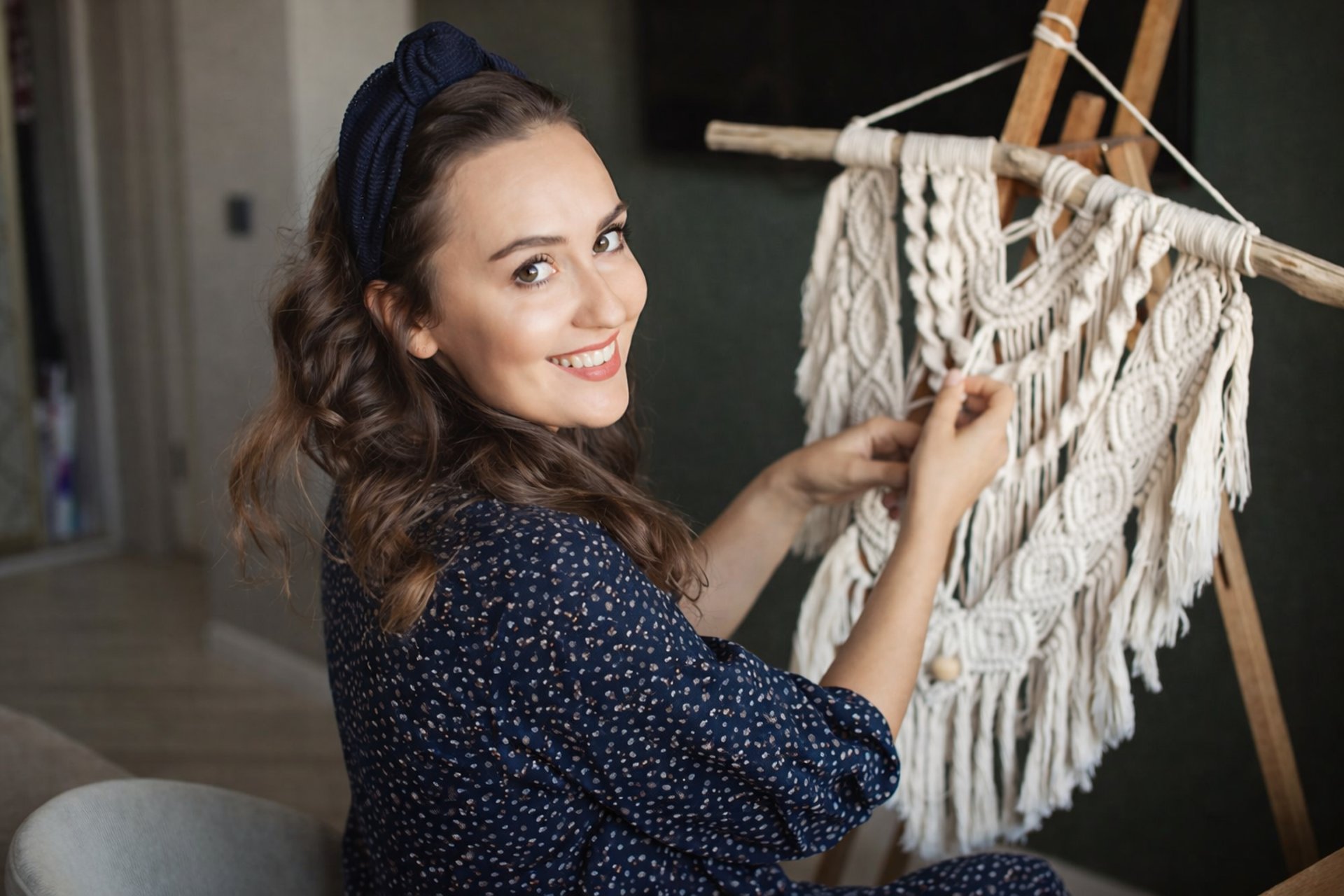 Artesana trabajando un tapiz de macramé hecho a mano, sonriendo mientras ajusta los nudos en un soporte de madera.