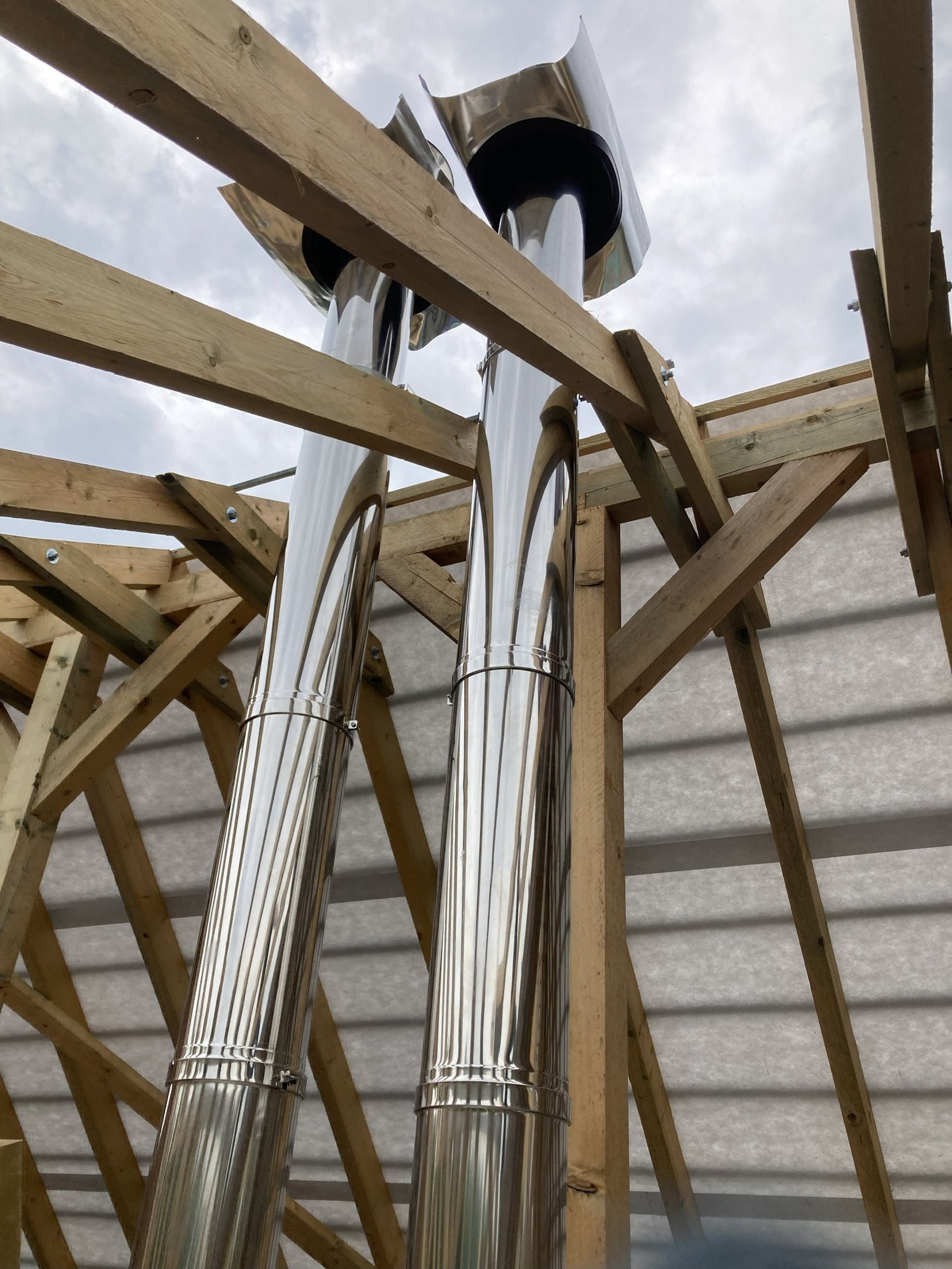 Két, korrózióálló acélból készült, fényes felületű szerelt kémény egymás mellett, egy épülő családi ház tetőterében.