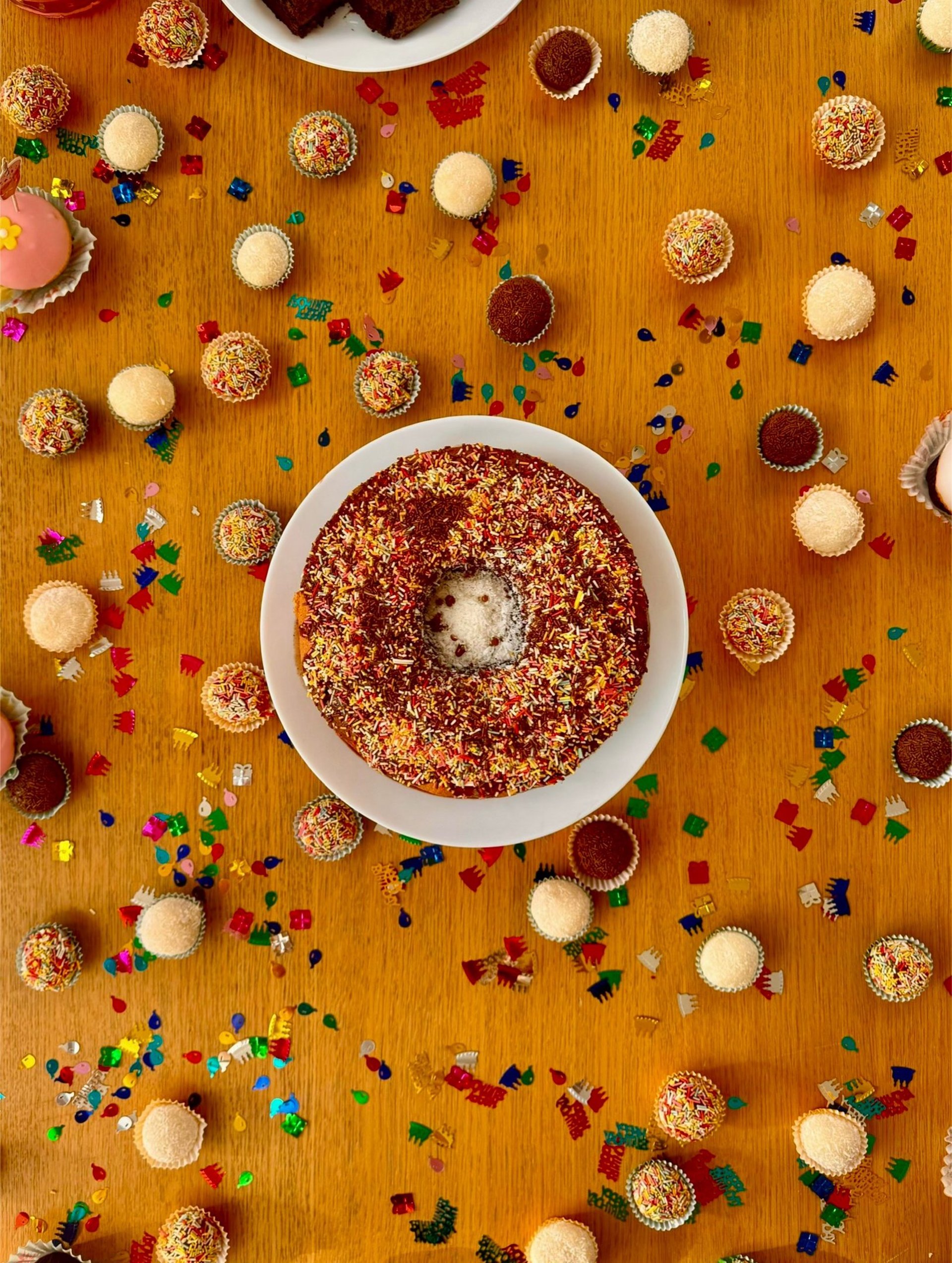 Donut-shaped cake topped with chocolate and sprinkles, surrounded by assorted brigadeiros and confetti on a wooden table.