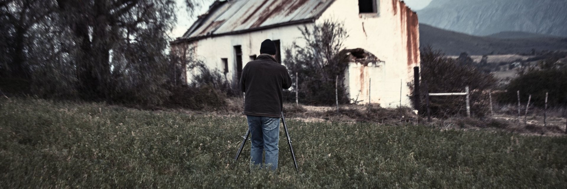 Martin Osner photographing a rural landscape in front of a weathered farmhouse with mountain scenery in the background.