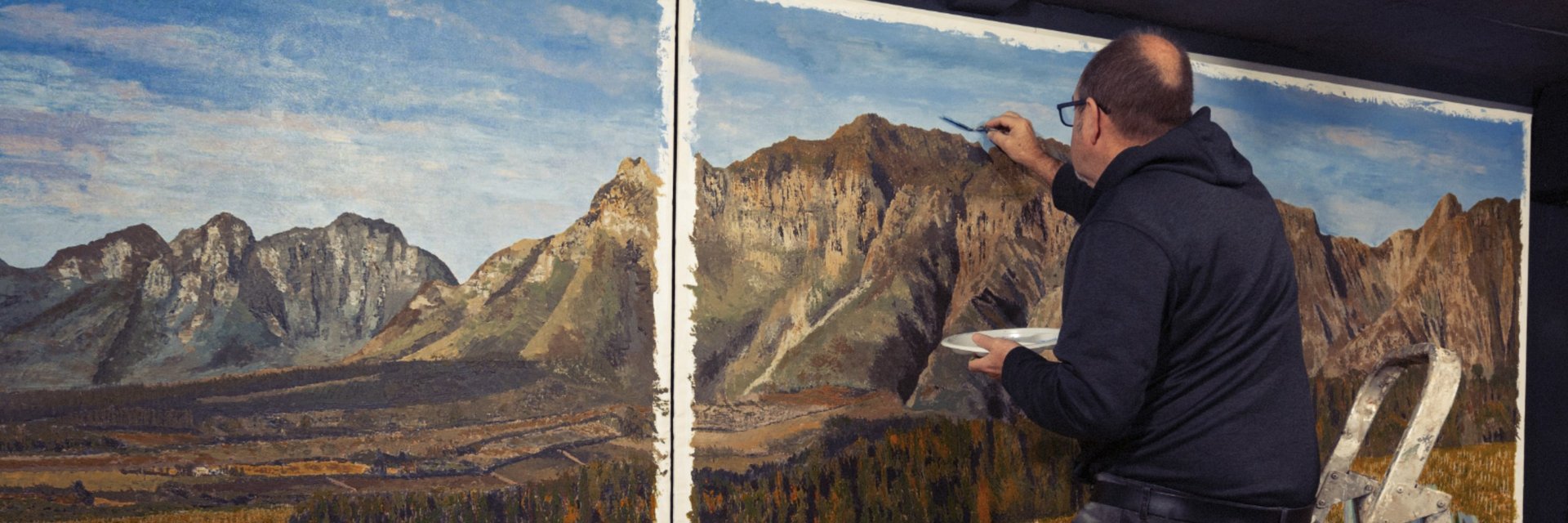 Martin Osner painting a large landscape artwork of mountains using a palette knife in his studio.