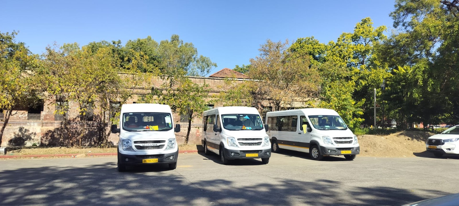 Three modern white Force Urbania vans parked in a sunny outdoor lot with trees and historic buildings in Delhi NCR