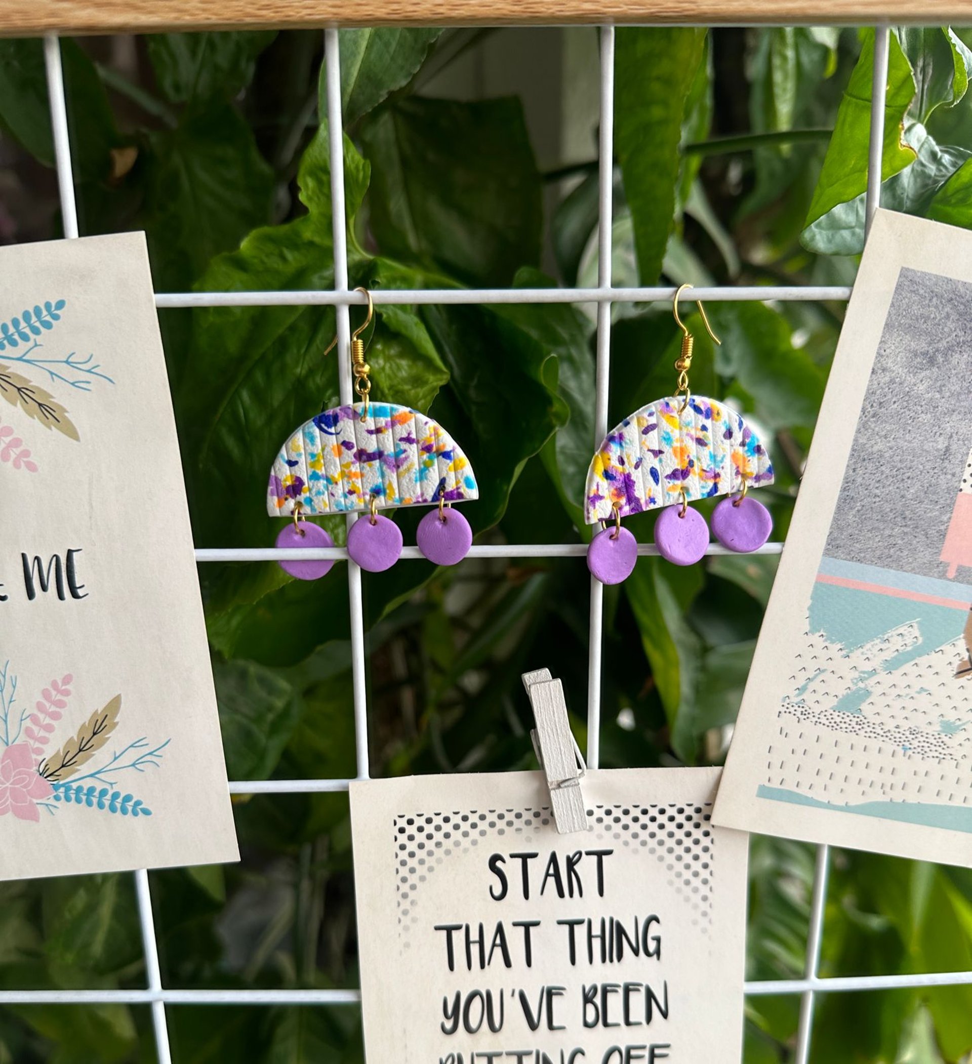 a pair of half-moon shaped colorful clay earrings hanging on wooden frame against leafy backdrop