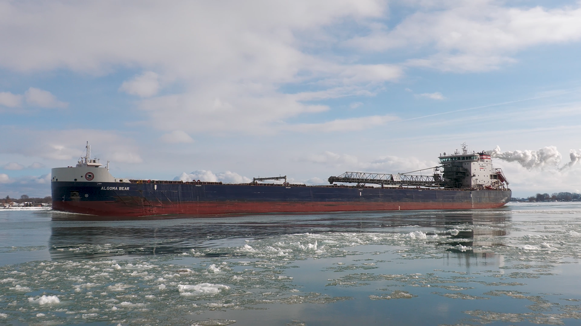 Freighter Algoma Bear from the front left side looking aft, on icy water with a plume of steam coming out of her funnel.