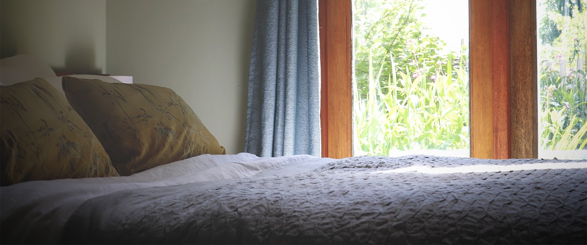 Double bed with mustard covered pillows and a grey cover with French doors overlooking a lush garden