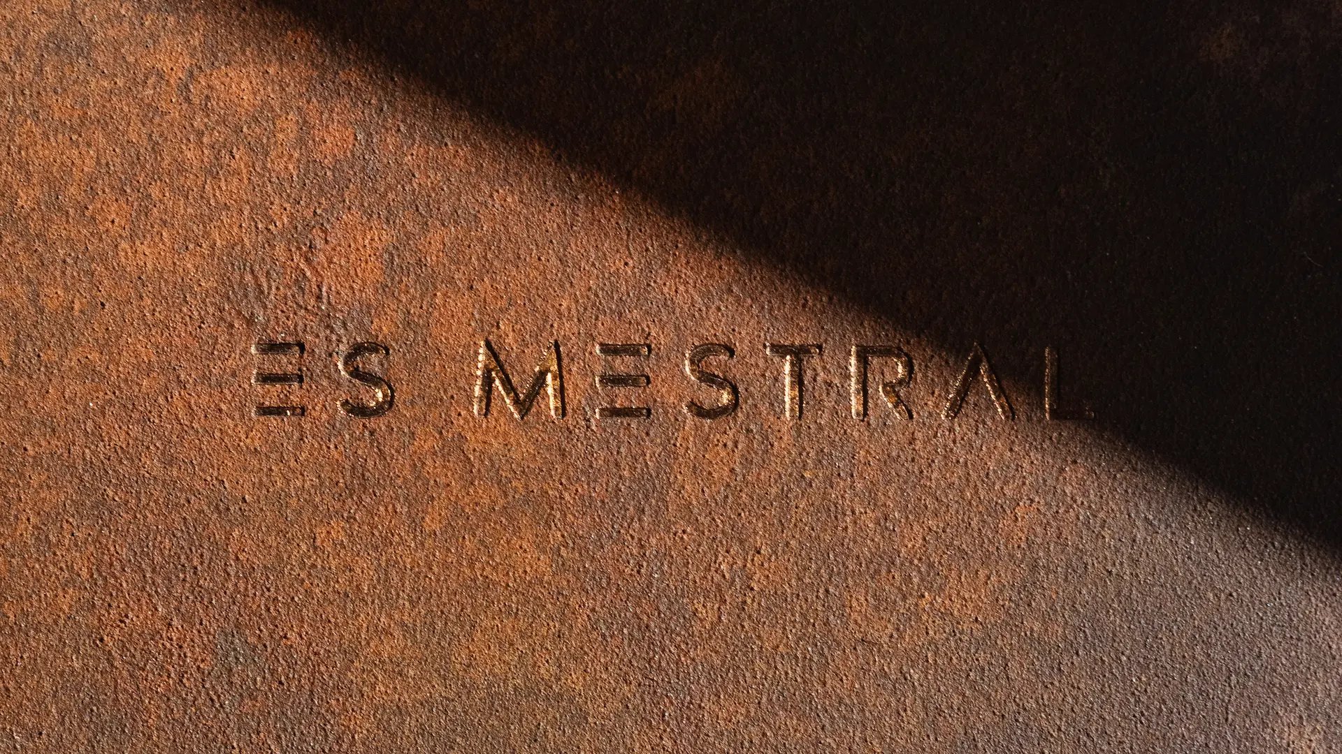 Detalle del logotipo Es Mestral grabado sobre superficie metálica oxidada, con luz y sombra que acentúan su textura.