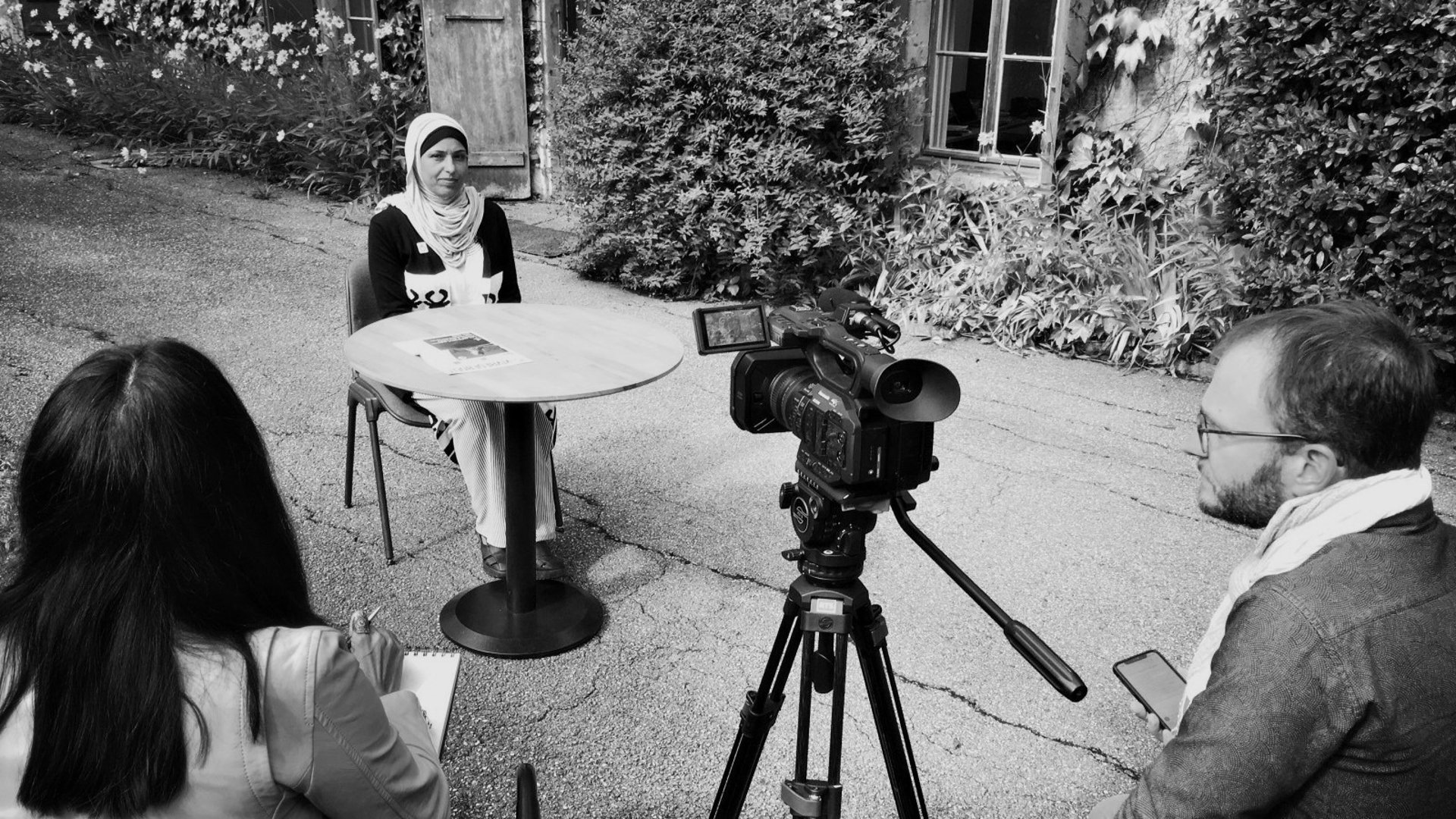A woman in a hijab sits at a table outdoors facing a video camera on a tripod, with two other people partially visible.