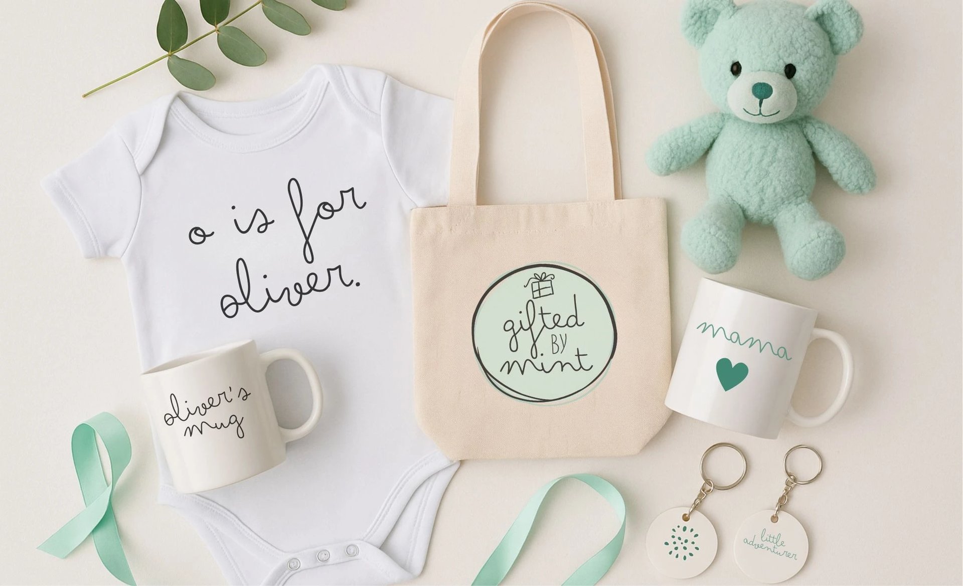 Flat lay of personalised baby gift set from Gifted by Mint including a white baby onesie with text ‘o is for Oliver’, beige t