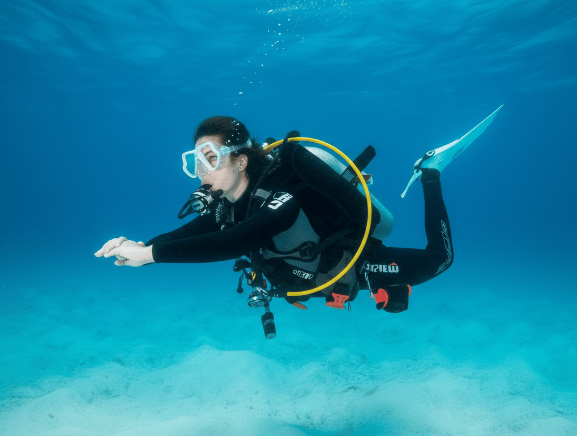 A female scuba diver swimming horizontally over a white sandy seabed in clear, bright blue tropical water.