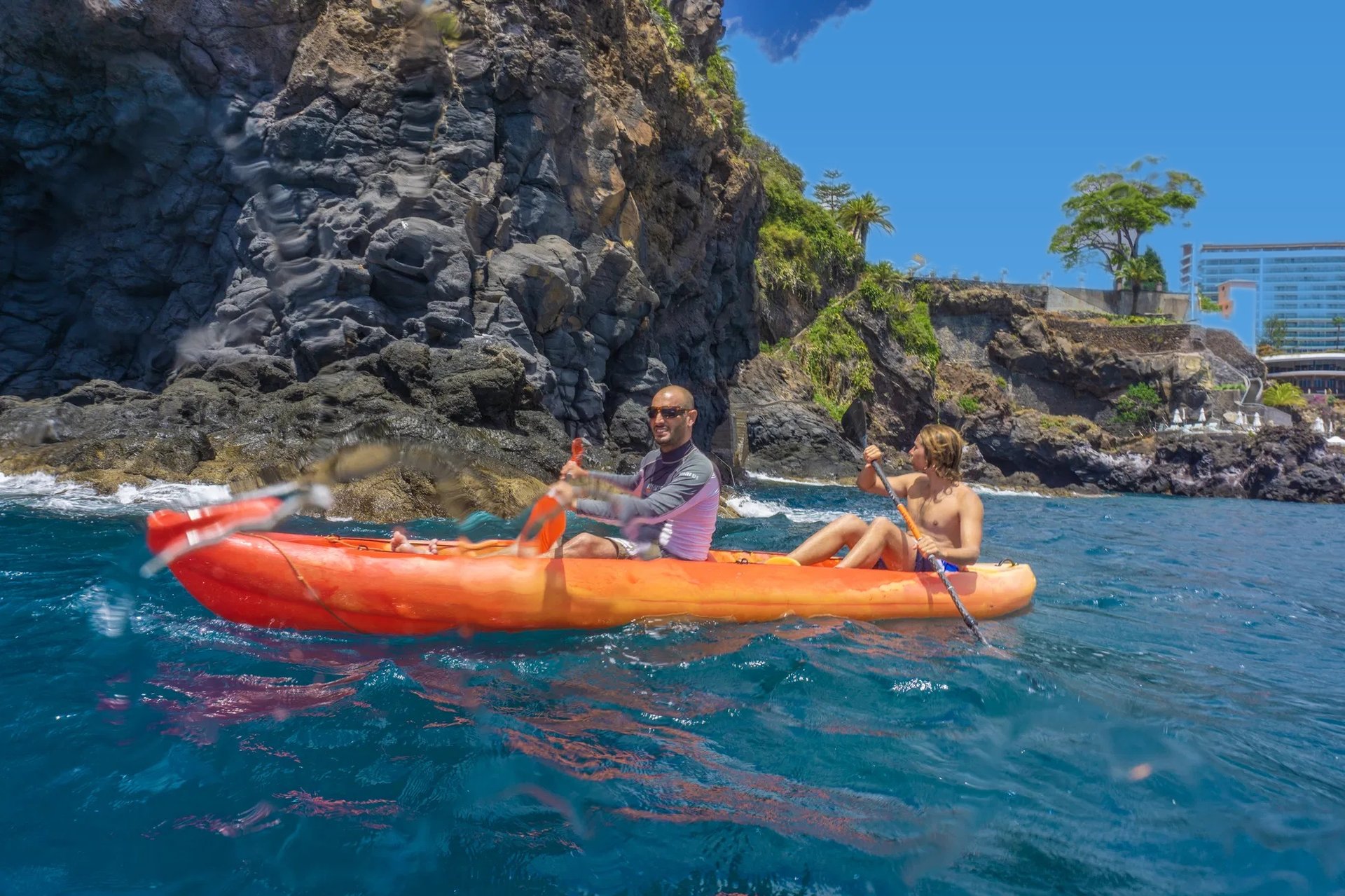 Guide and guest paddling an orange tandem kayak along the rocky volcanic cliffs of Funchal, Madeira