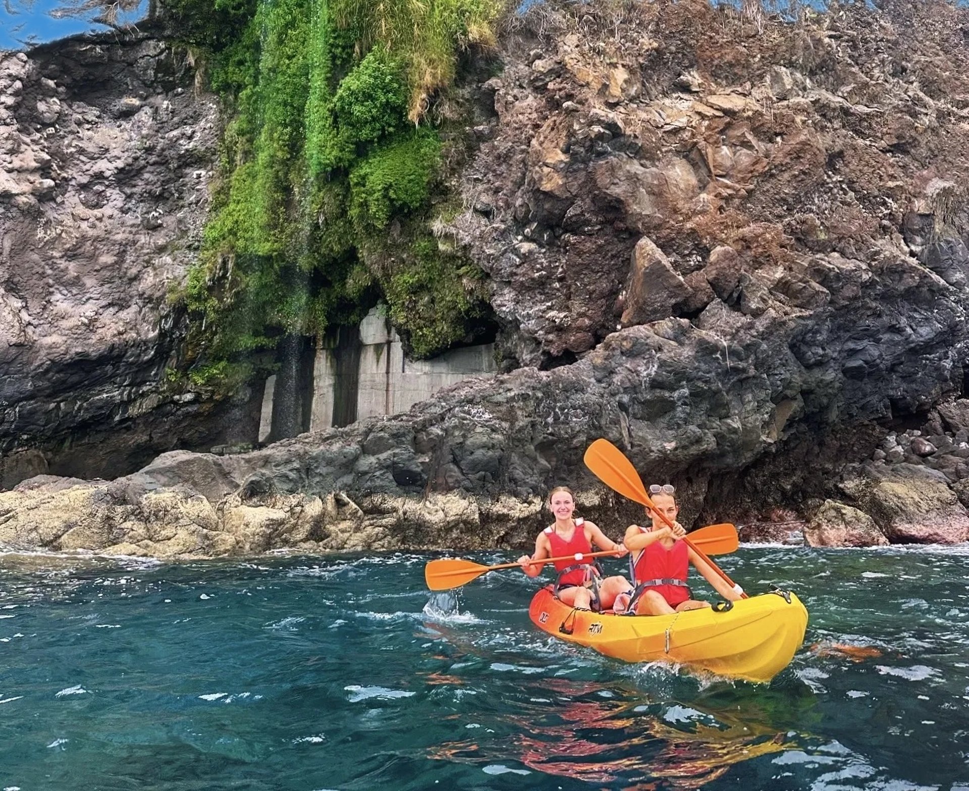 Two women in lifejackets paddling a yellow kayak past a green cliff waterfall on the Madeira coast