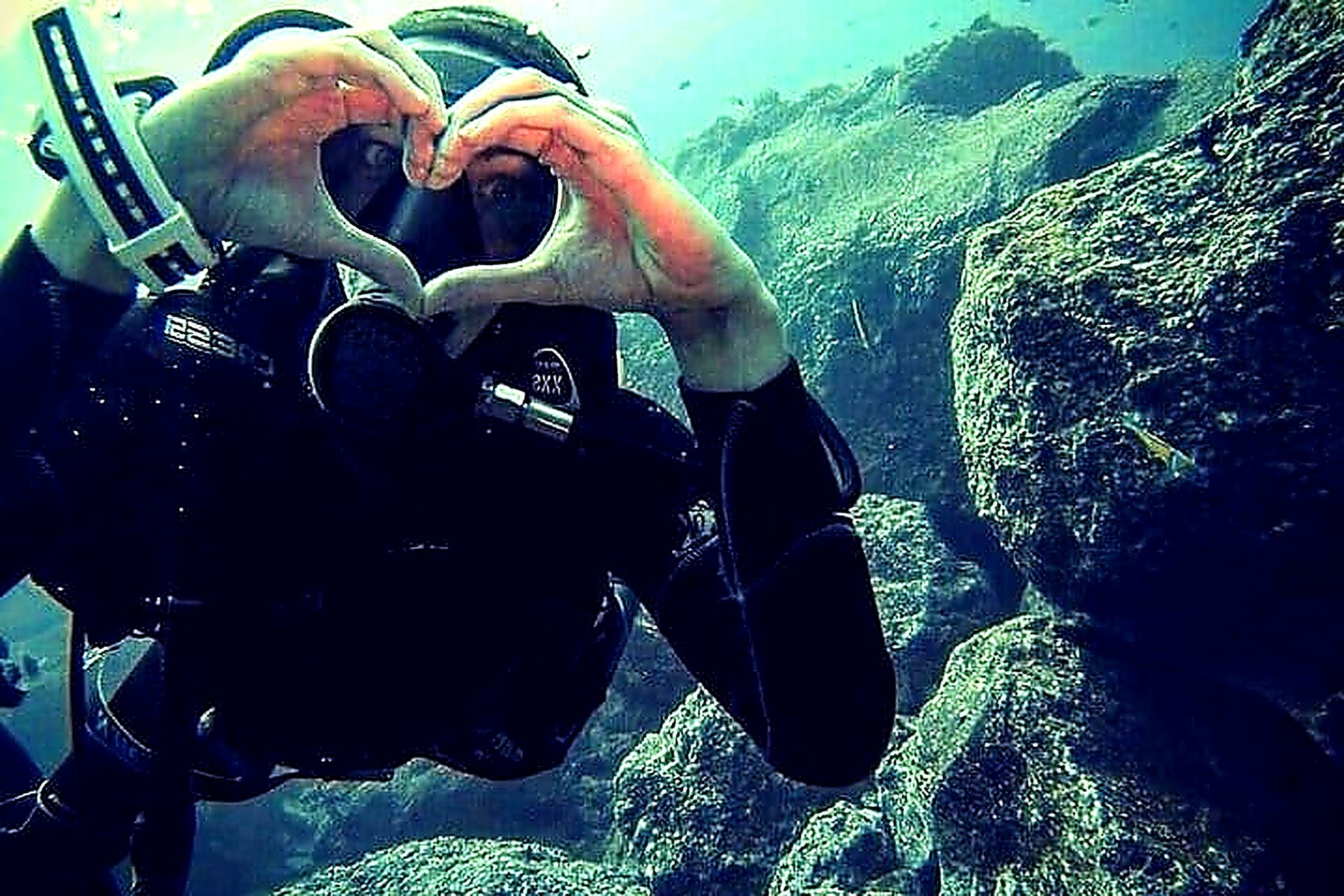 Scuba diver making a heart shape with hands underwater among rocks in Funchal, Madeira