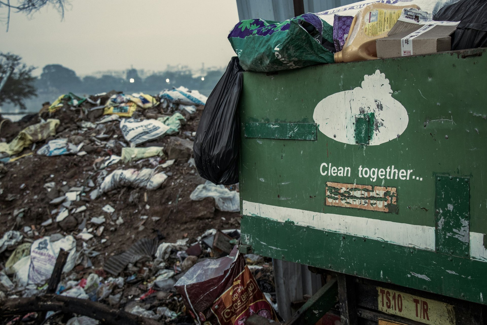 Grön sopcontainer med texten ”Clean together…” och svarta sopsäckar, framför skräpig tipphög - visar behovet av regelbunden s
