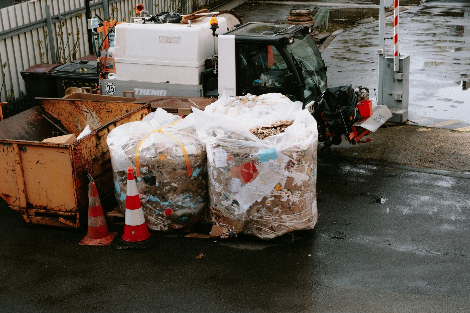 Två storsäckar med blandat pappersavfall på våt gård bredvid rostig container, varningskoner och servicefordon - industriell 