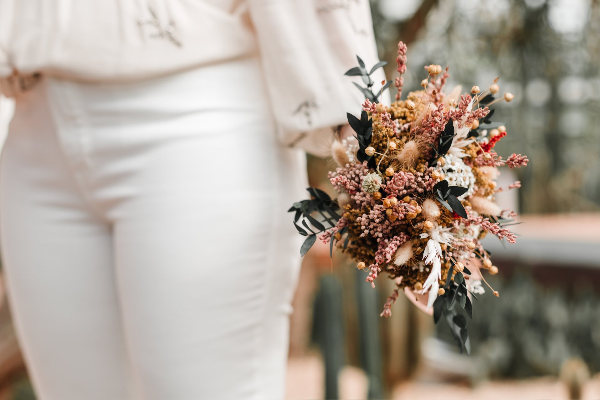 Primer plano de la mano de una novia que sostiene un ramo de novia de estilo bohemio con flores secas y follaje texturizado.