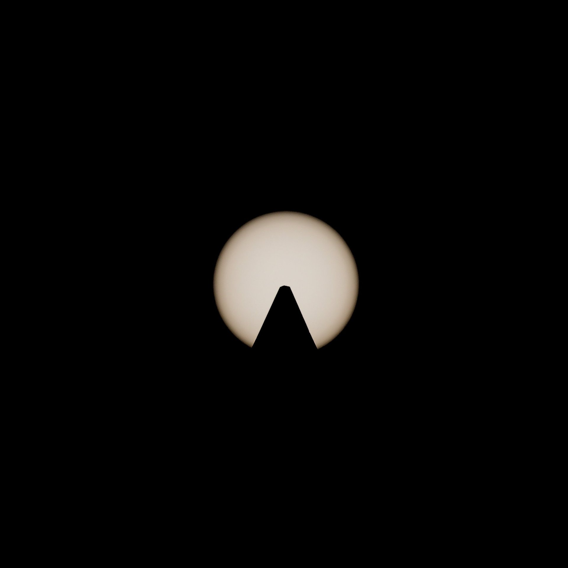 Symmetrical silhouette of an obelisk peak centered against a glowing sun, captured with a solar filter.