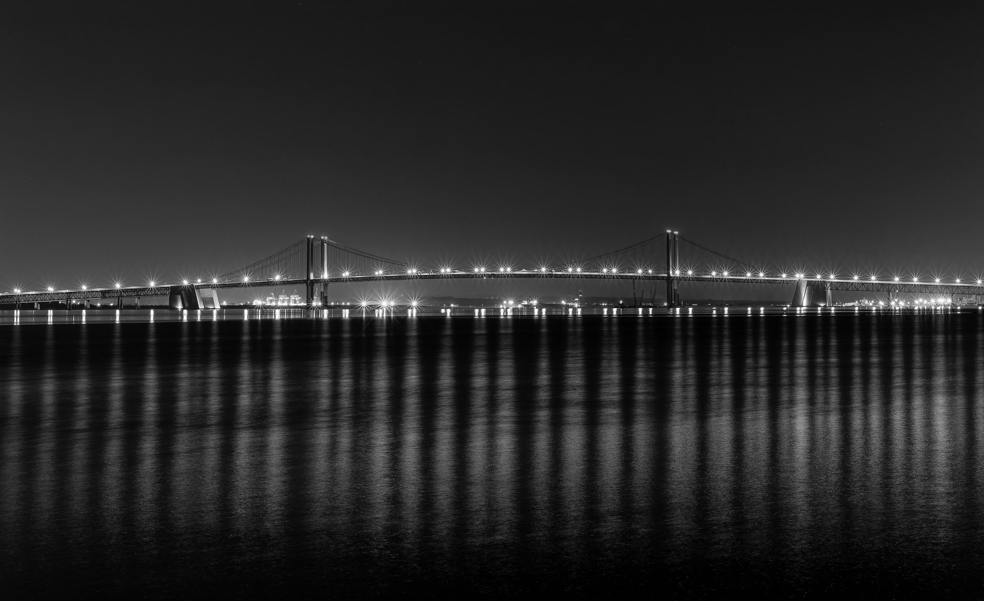 Black and white photo of the Delaware Memorial Bridge at night; glowing lights create rhythmic vertical reflections.