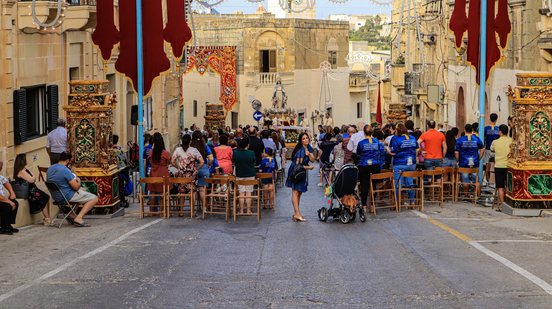 People sitting in a narrow street in front of a statue of St Mary.