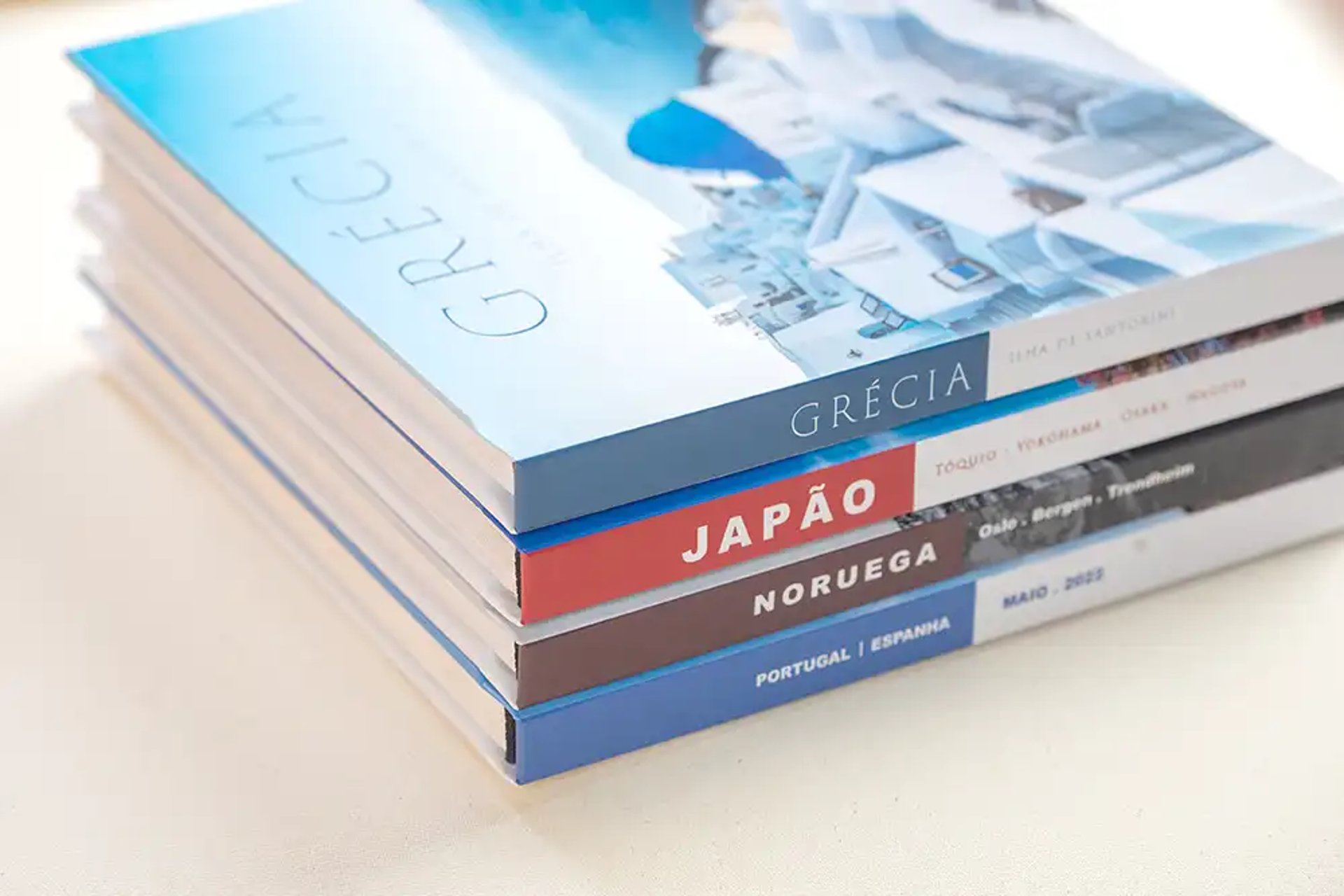 Pilha de fotolivros personalizados com capas temáticas de viagens para destinos como Grécia, Japão, Noruega e Portugal.