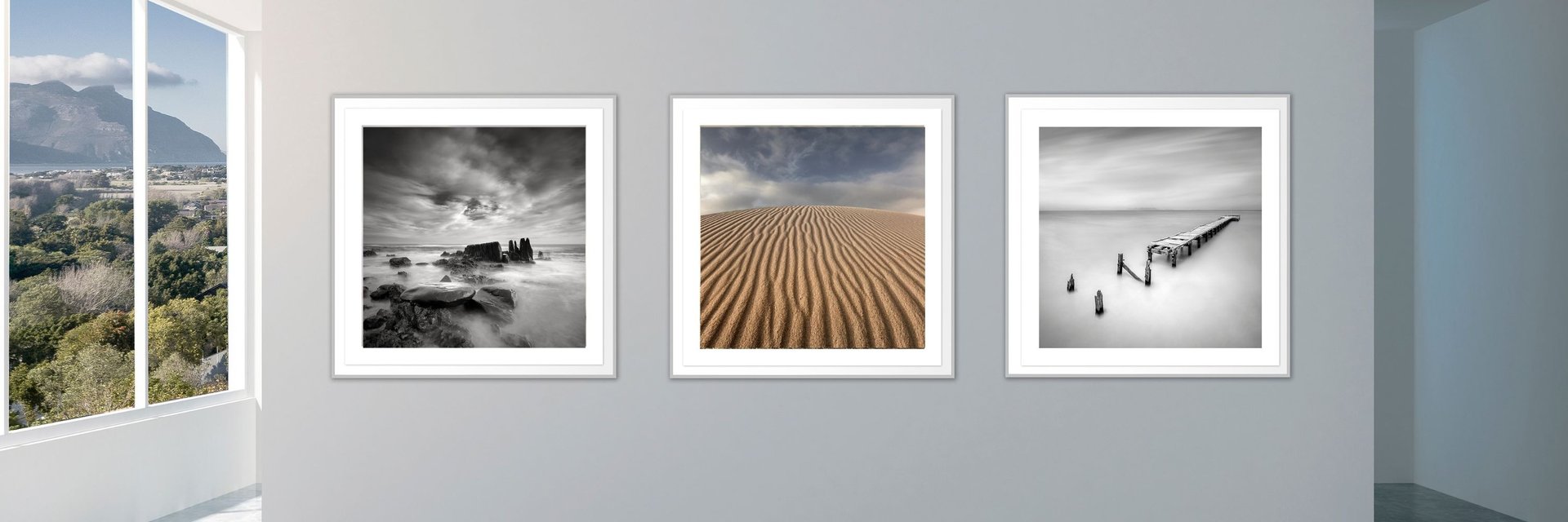 Framed fine art landscape photographs displayed on a modern gallery wall with mountain views through large windows.