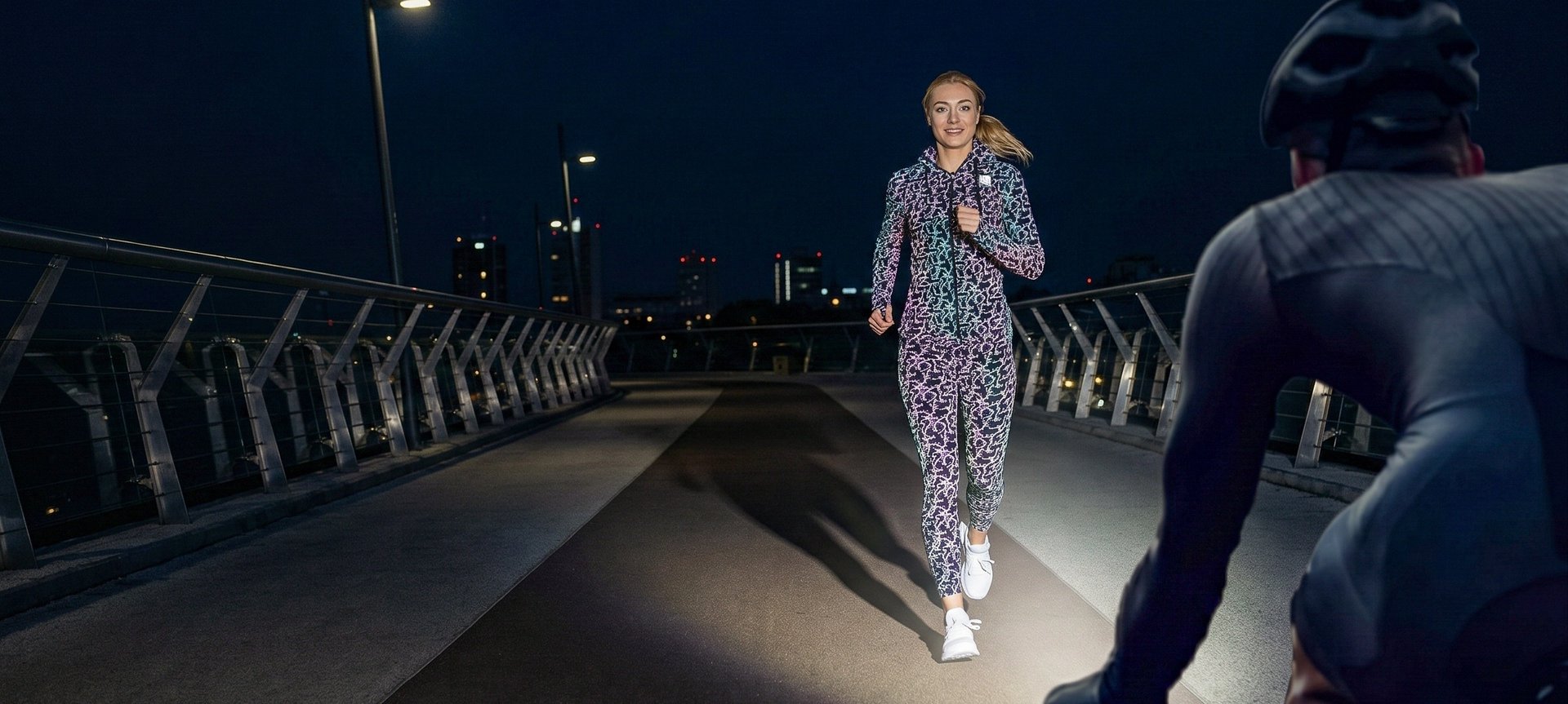 Wide shot of a woman running at night on a bridge, highlighted by a cyclist's headlight