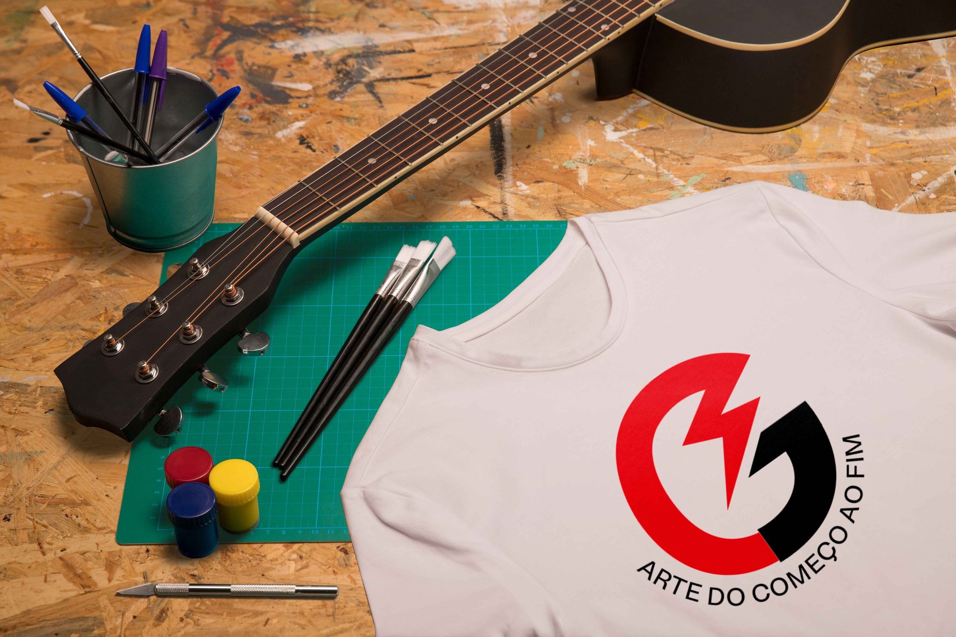 T-shirt estampada com o ícone da marca Garage sobre uma mesa onde estão tintas, pinceis e um violão