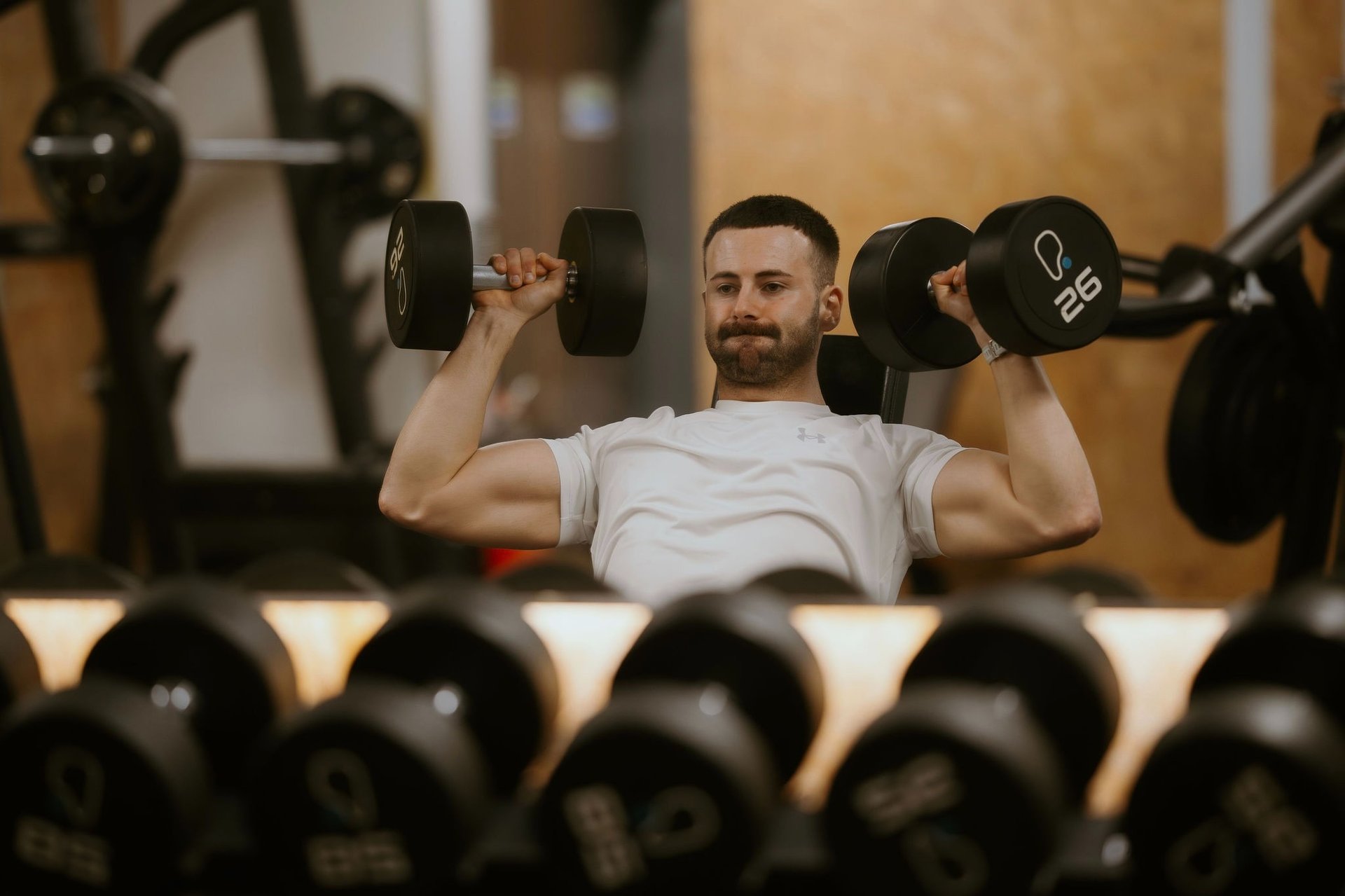 Trainer performing seated dumbbell shoulder press with 26kg weights in Leamington Spa gym, focused on strength and form