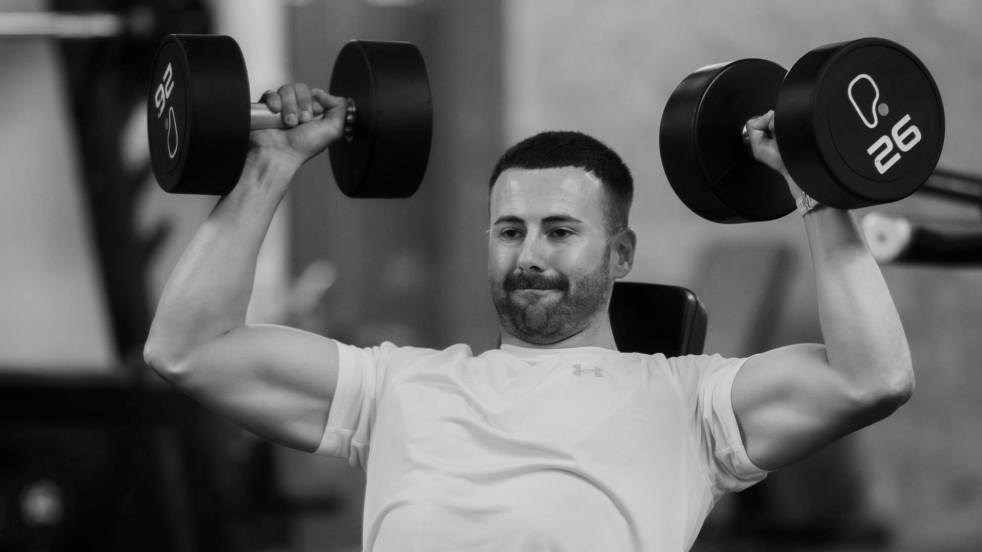 Trainer performing seated dumbbell shoulder press with 26kg weights in Leamington Spa gym, focused on strength and form