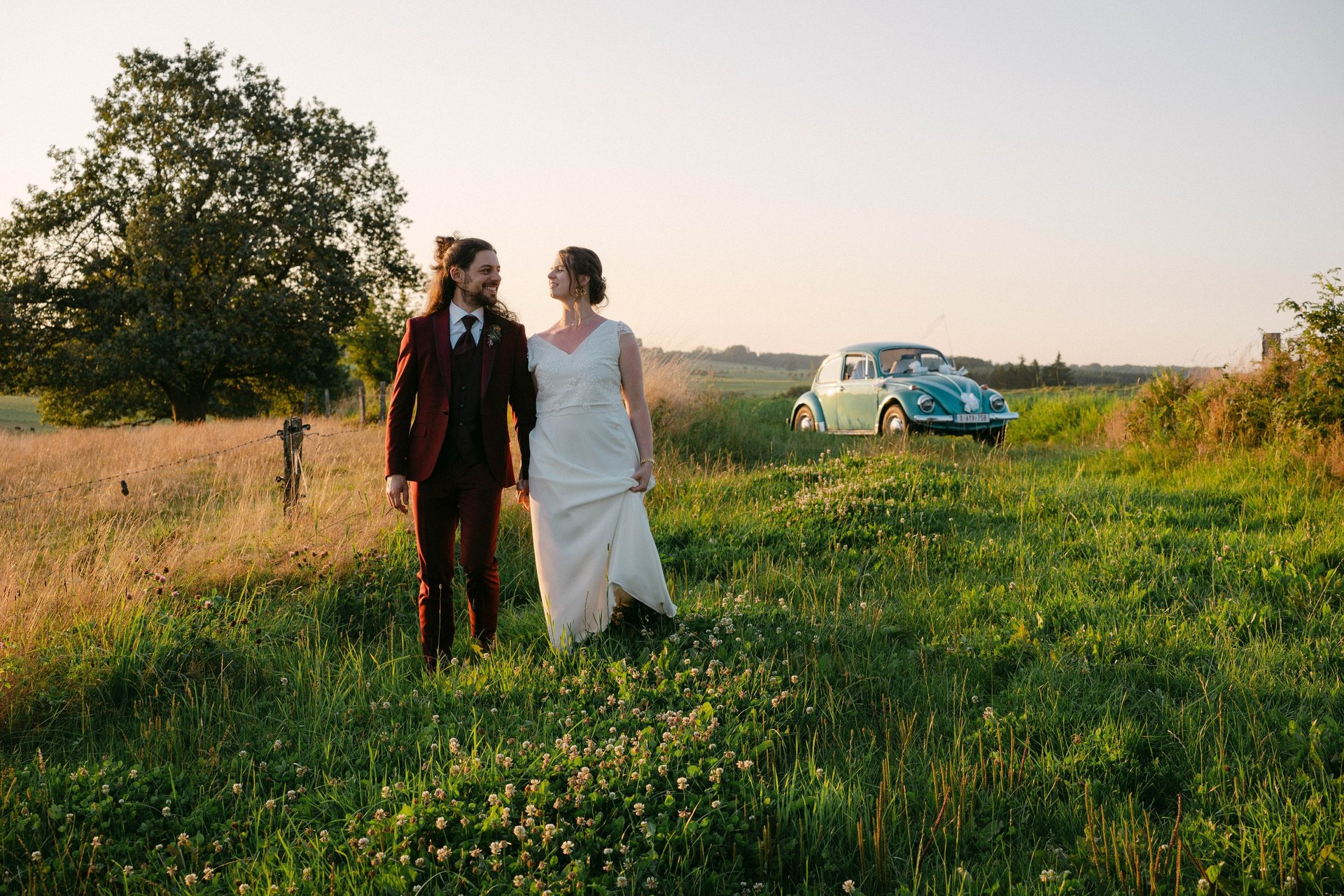 Couple de mariés marchant dans un prré avec leur old timer (une vw coccinelle ) derrière eux