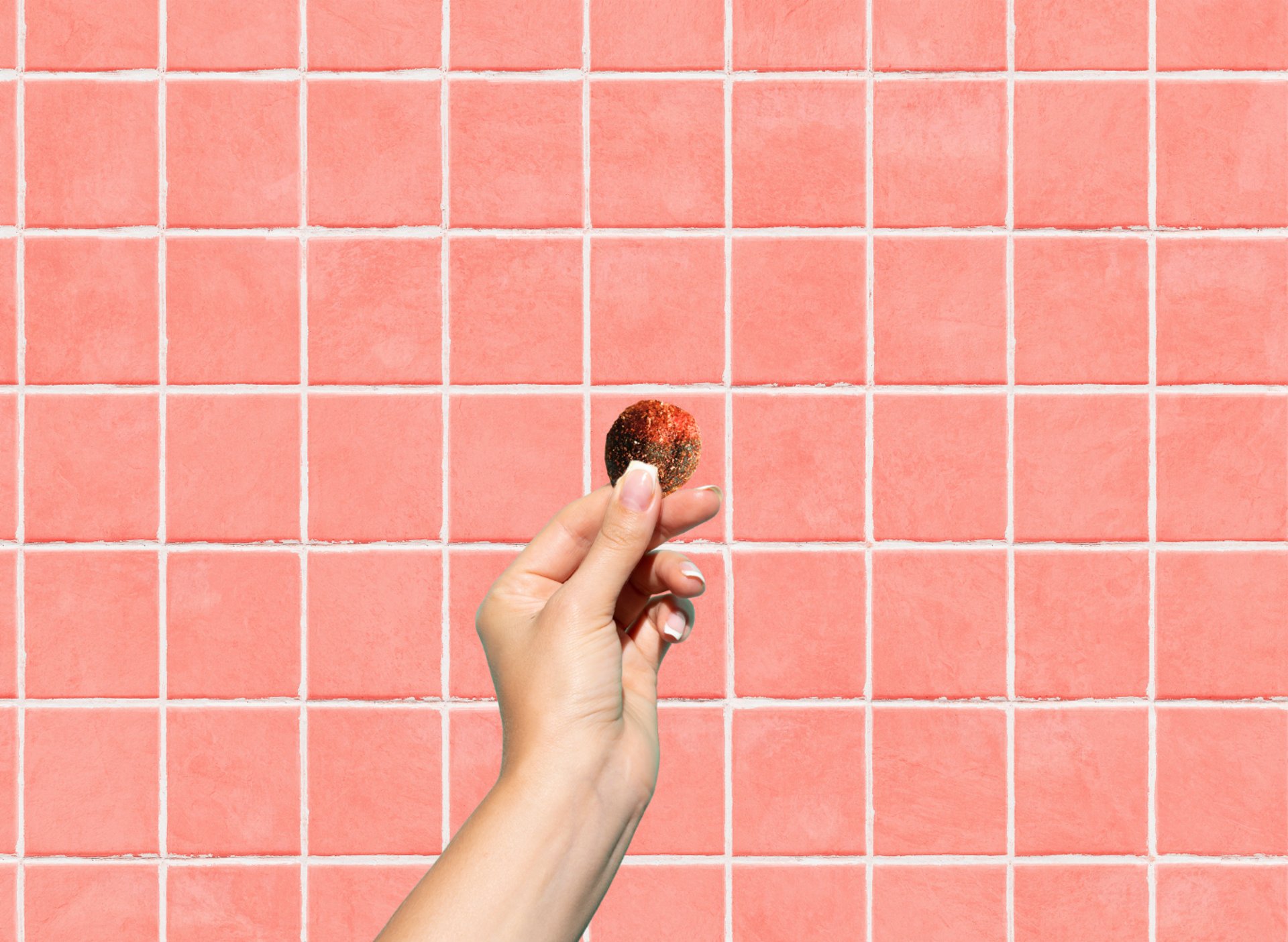 A hand holding a round piece of gummy against a pink tiled wall background.