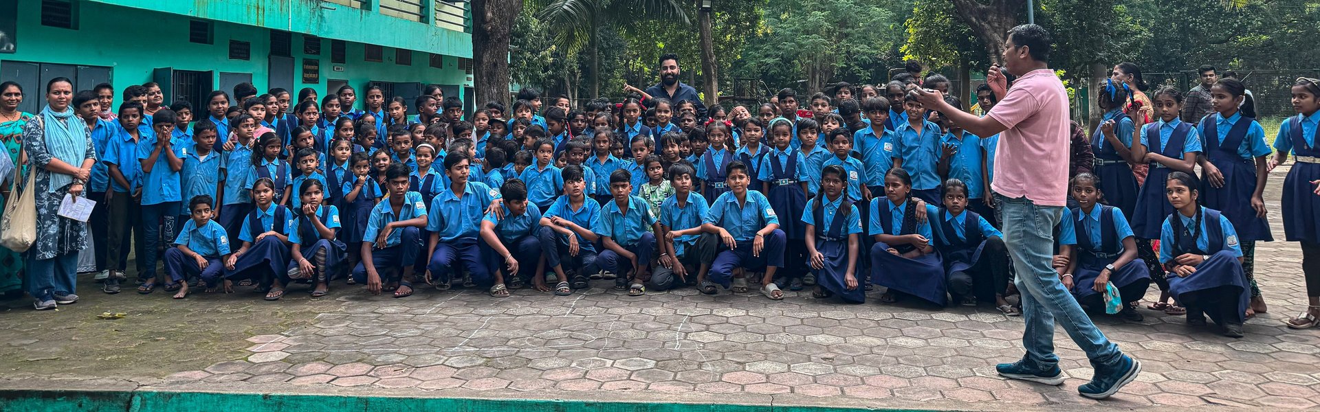 Educator addressing a large group of students in blue uniforms gathered outdoors at a school campus