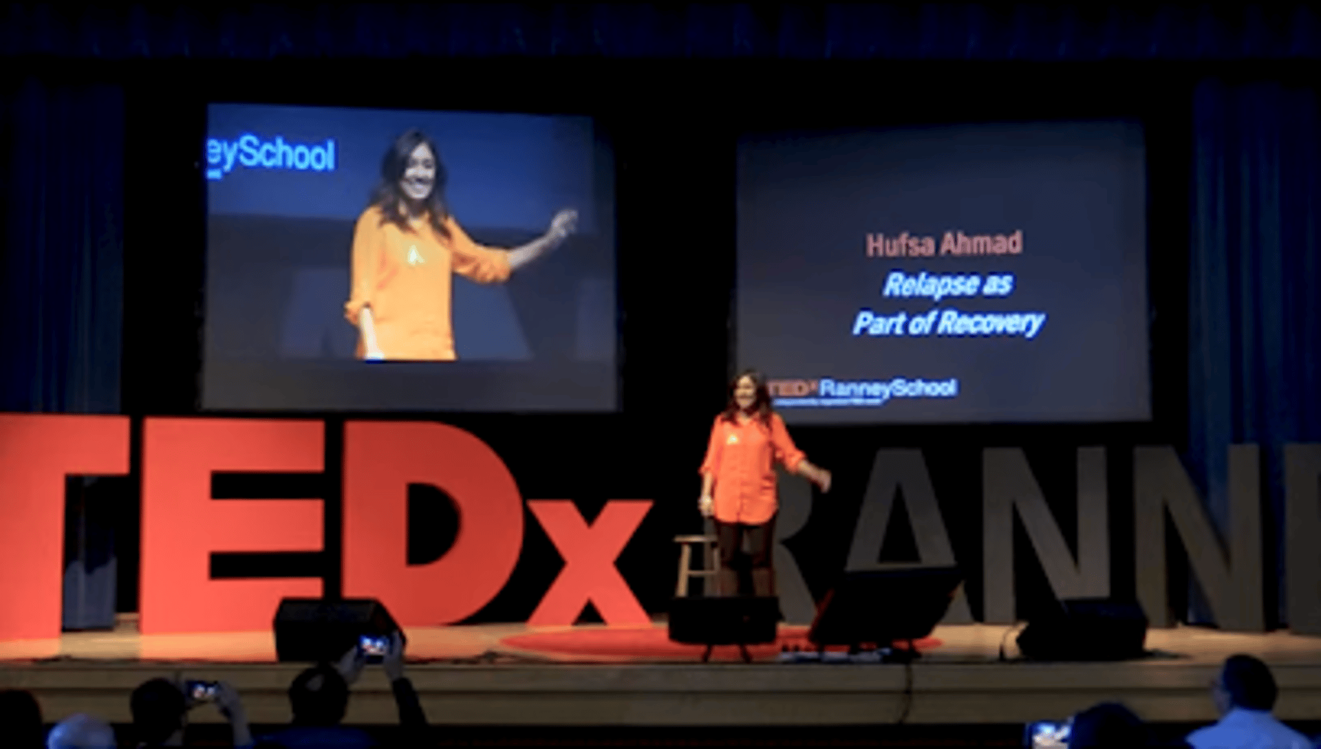 A woman in an orange shirt smiles on stage, wearing a green awareness ribbon, conveying positivity.