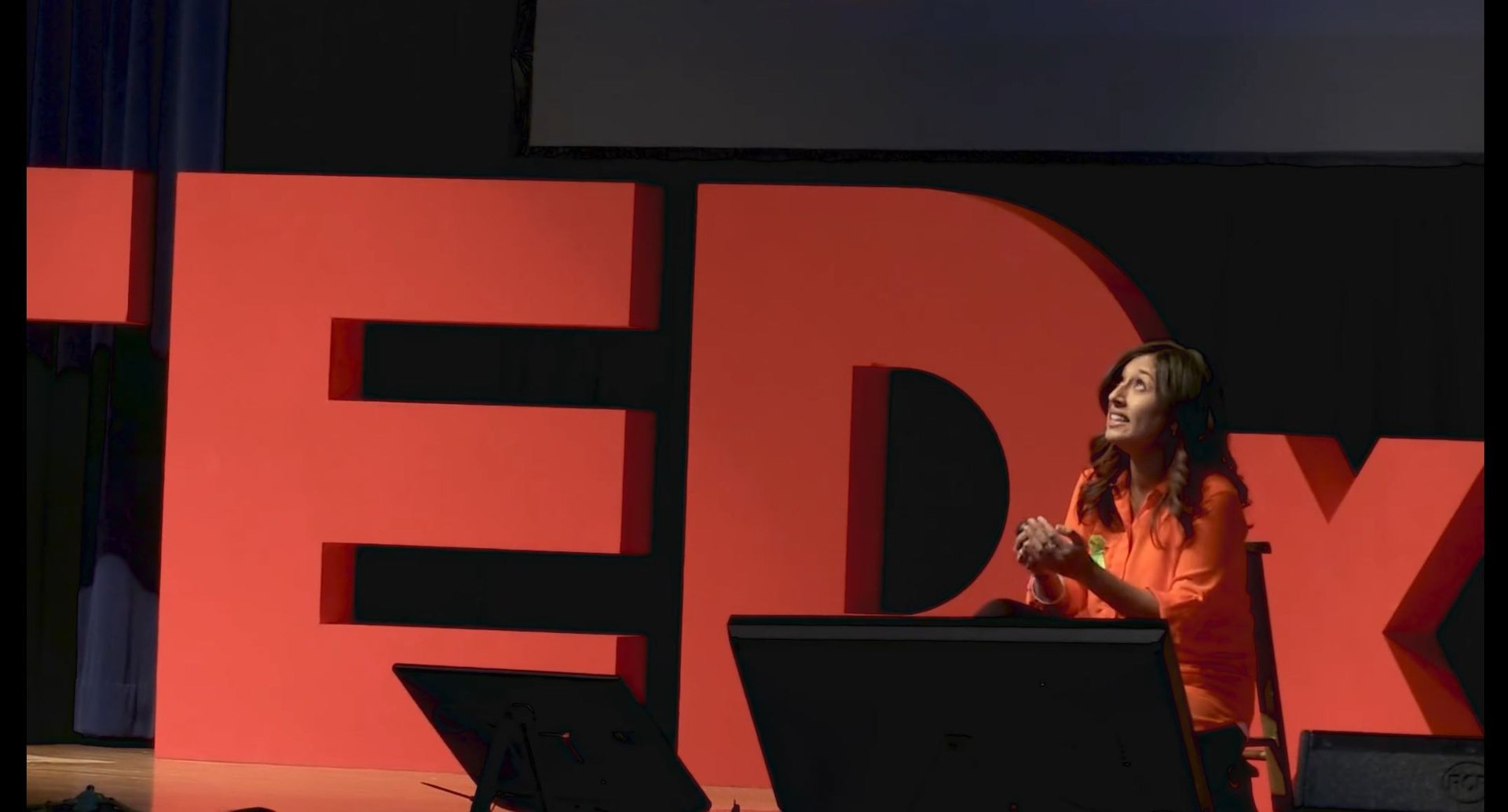 A woman in an orange outfit speaks passionately on stage, with large red "TEDx" letters behind her.