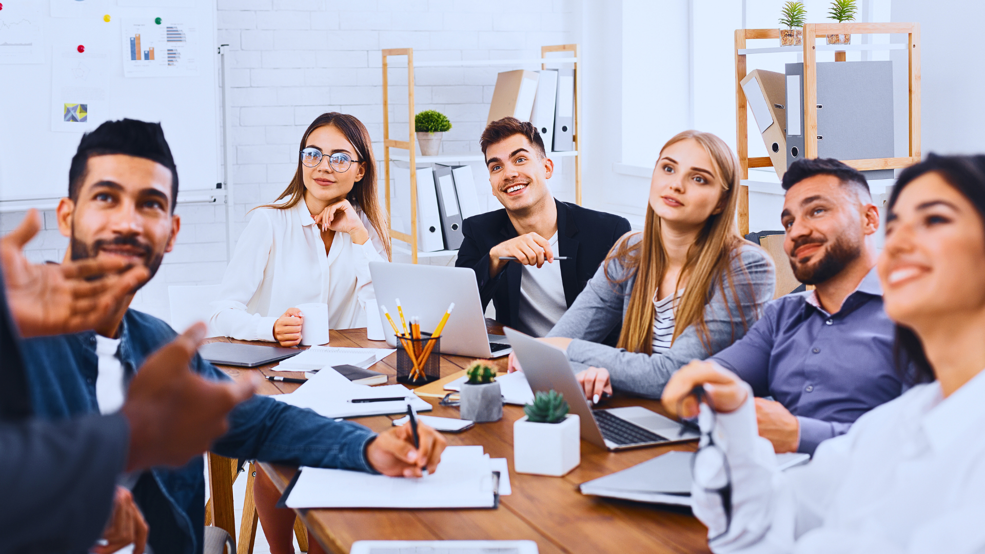 Group of business leaders with excited expressions sit at a table, all looking up at a standing presenter who is offscreen.