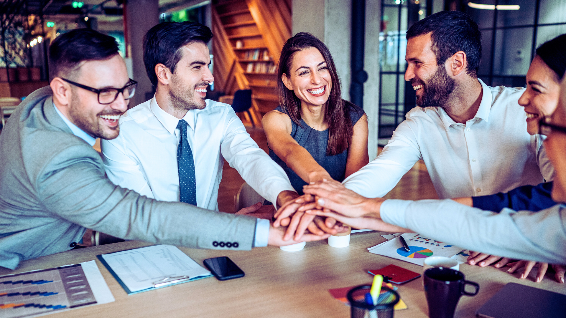Group of business leaders sits around a table, participating in a team huddle, each extending their right arm to touch hands.