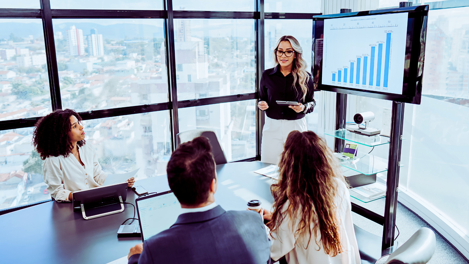 Woman in glasses and a black shirt presenting a bar chart on a TV screen to two women and a man in a glass-walled boardroom