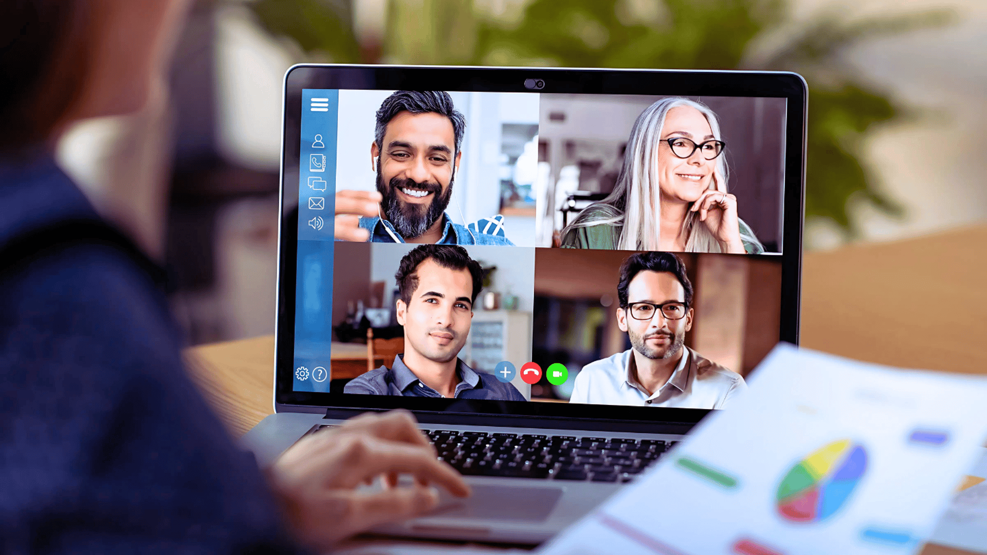 A business consultant holds printed graphs while hosting a virtual meeting with four people on their laptop.