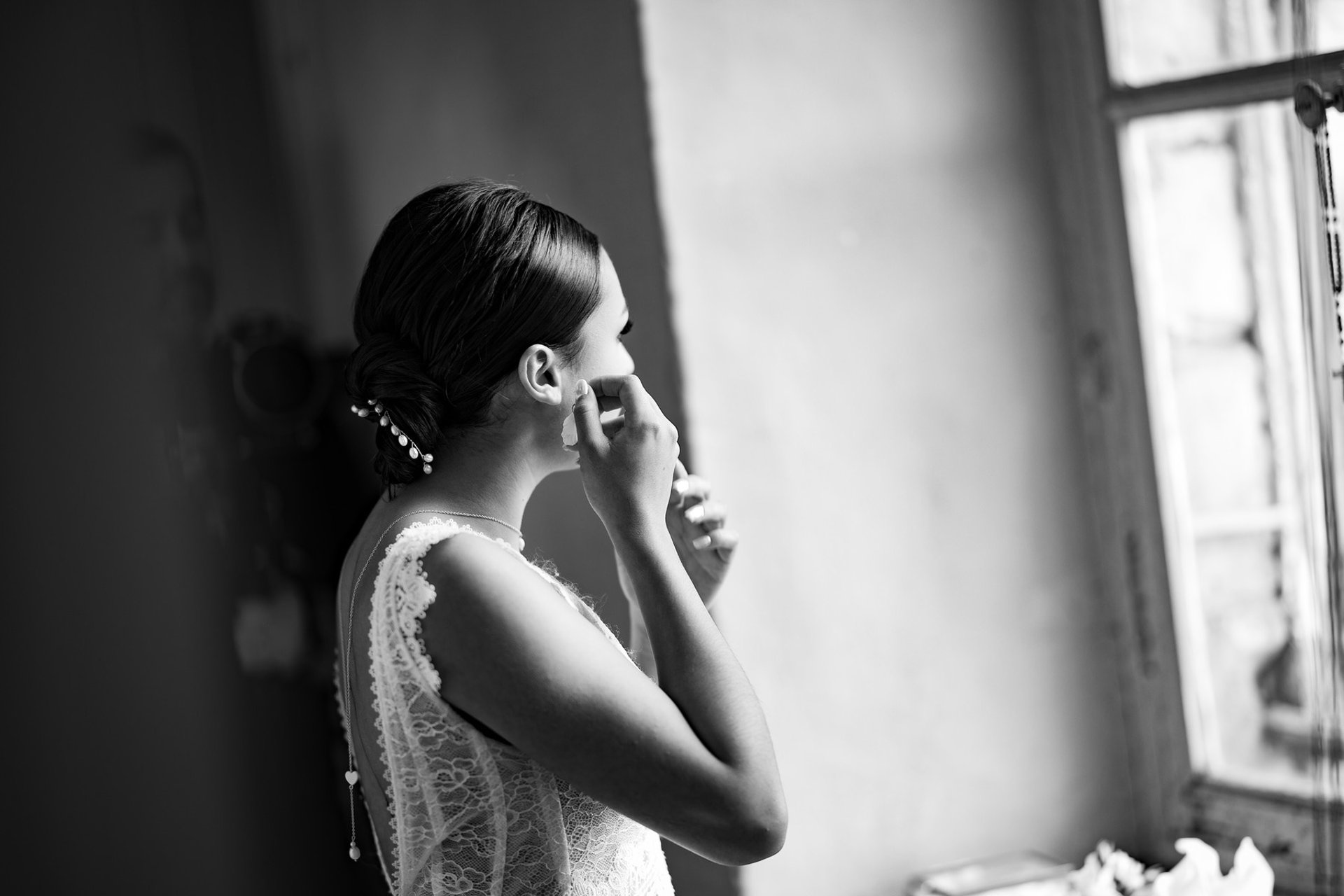 Une femme en robe de mariée accrochant sa boucle d'oreille face à une fenêtre