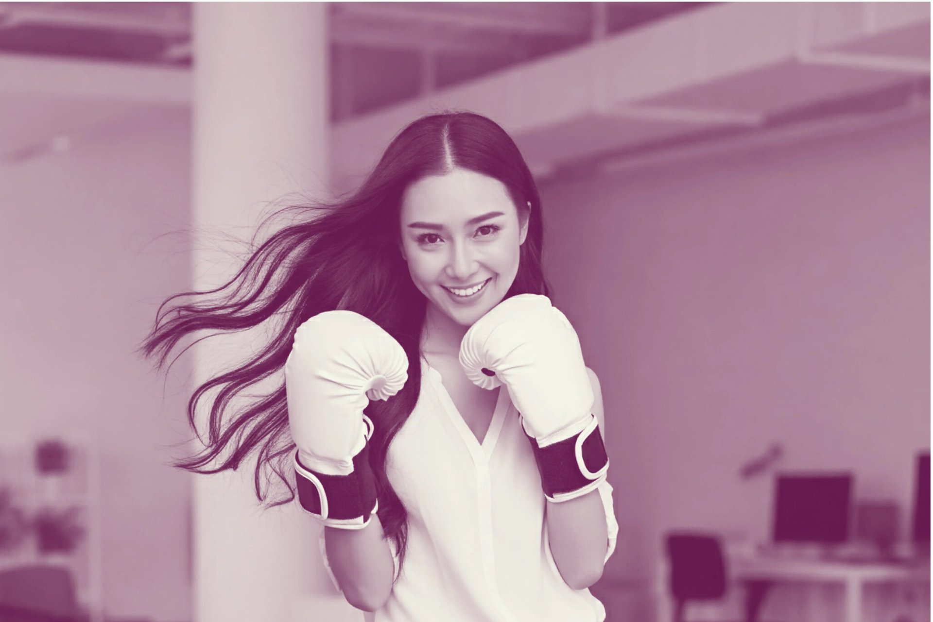 A smiling Asian woman in boxing gloves stands confidently in an office.