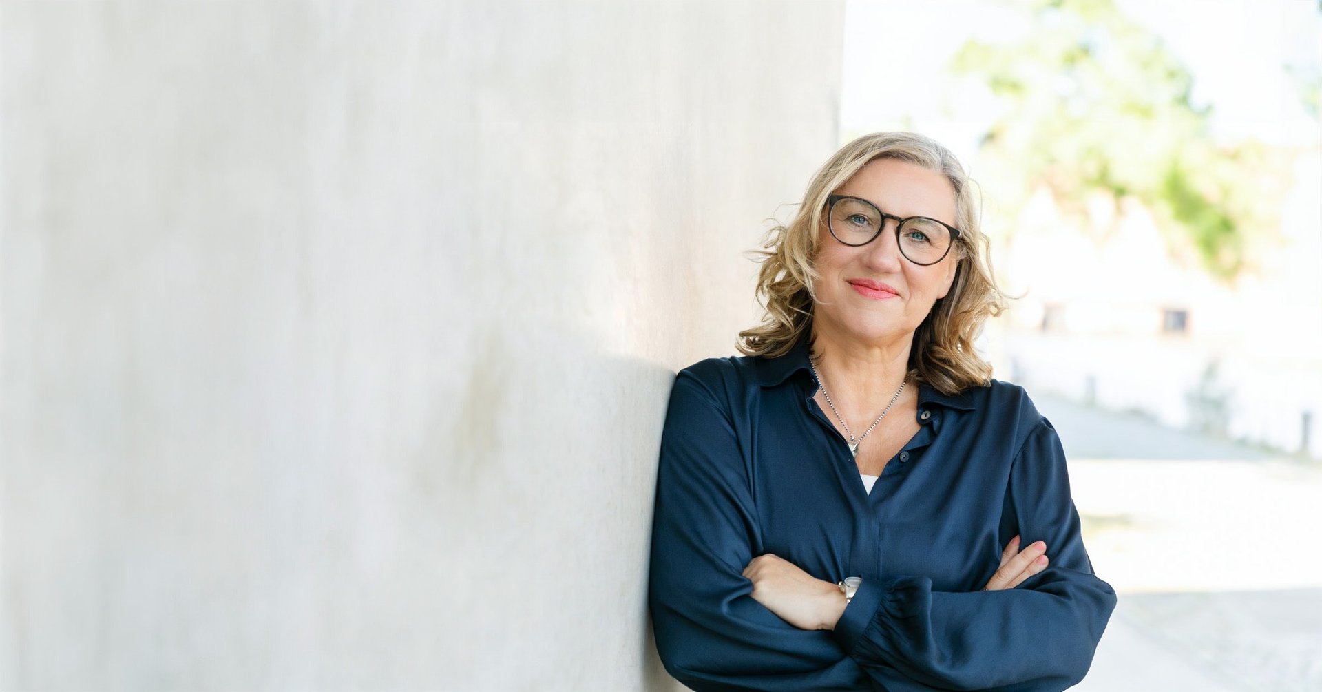 Gundula schaut freundlich lächelnd mit Brille in blauer Bluse angelehnt an einer hellen Betonwand.