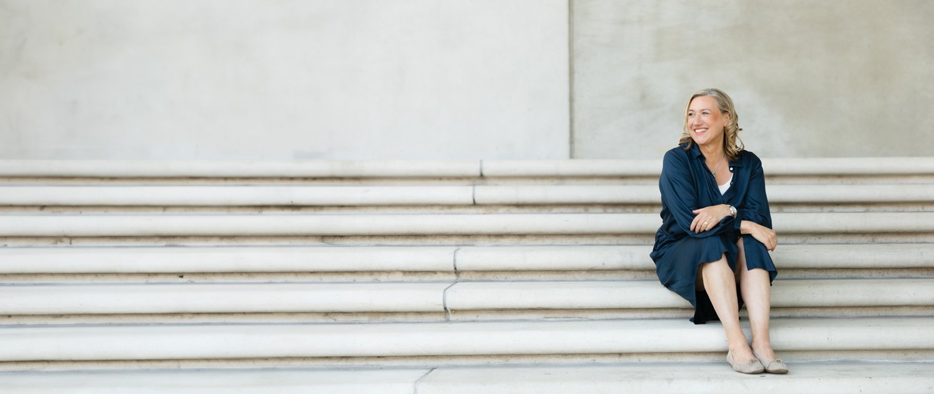 Gundula im blauen Kleid auf einer weißen Betontreppe sitzend.