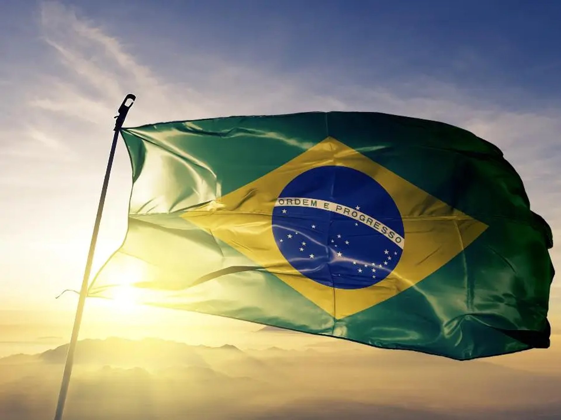 Imagem da Bandeira do Brasil, ao fundo o sol se pondo em uma montanha e sol ceu esta azulado com nuvens