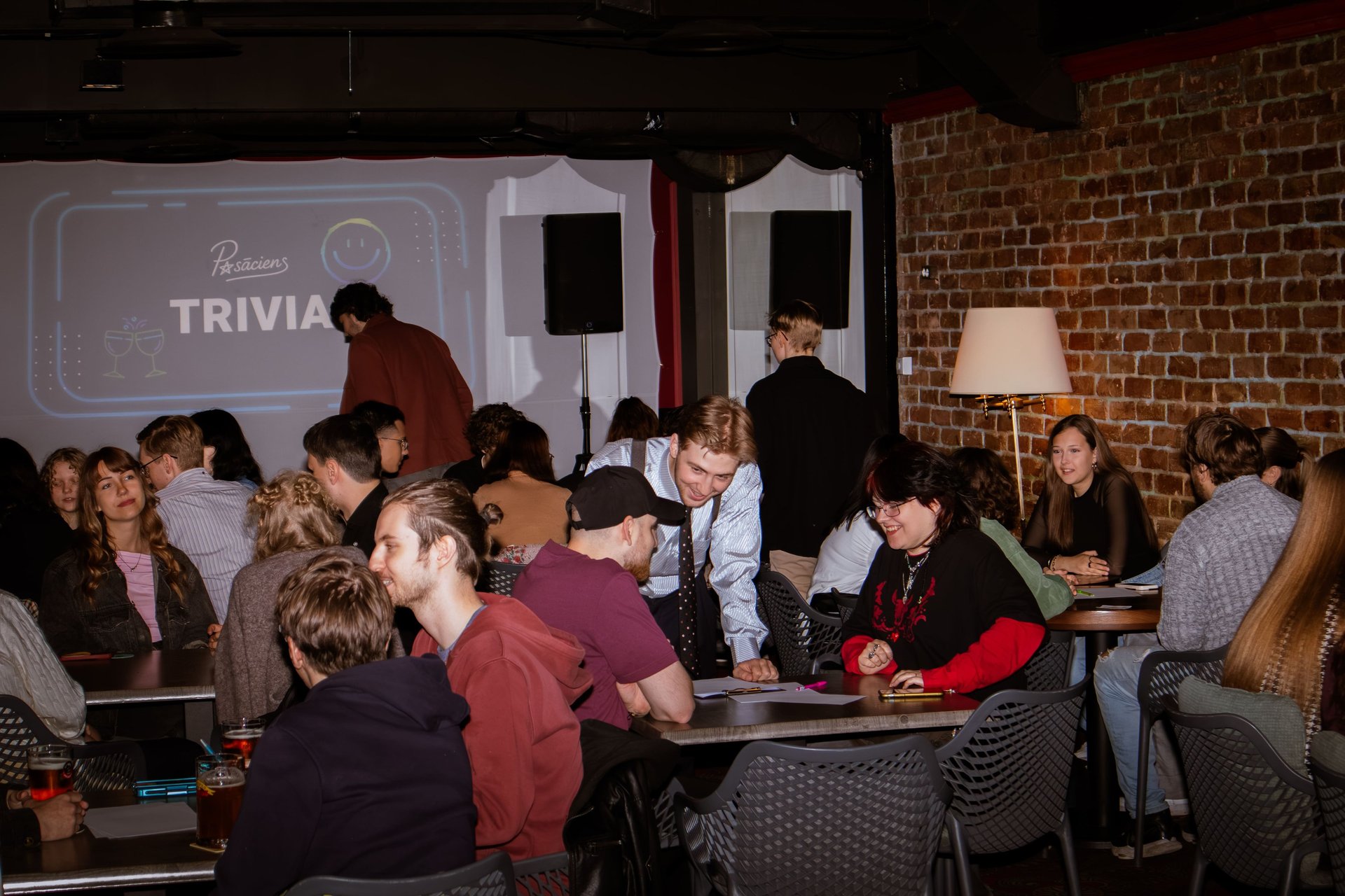 Viktorīna, jaunieši smaidīgi telpā, pasākuma organizatori un vadītājs pārvietojas pa telpu.