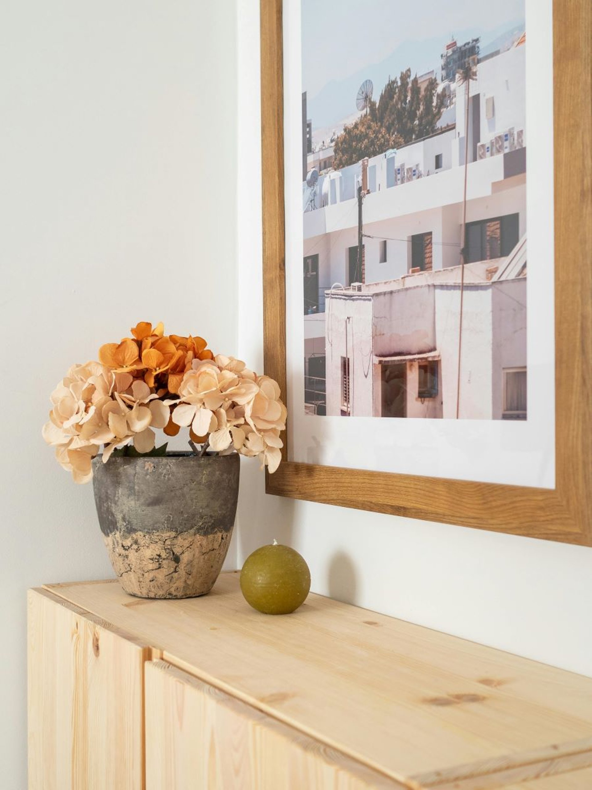 un pot d'hortensias posé sur un meuble en bois clair et un cadre avec des maisons au mur