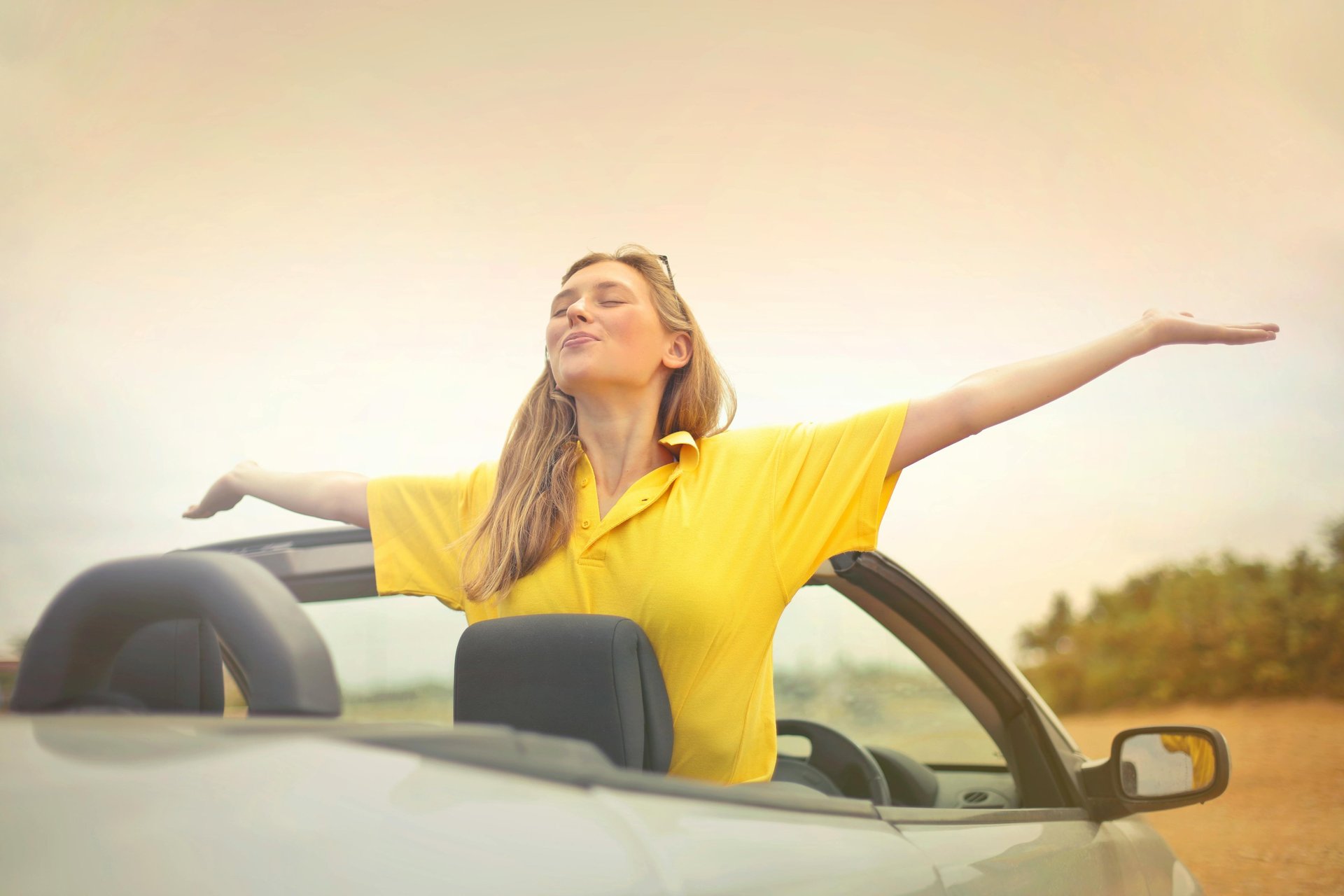 Woman in yellow t-shirt standing in a convertible car.The woman's eyes are closed,arms out basking in the sunshine.