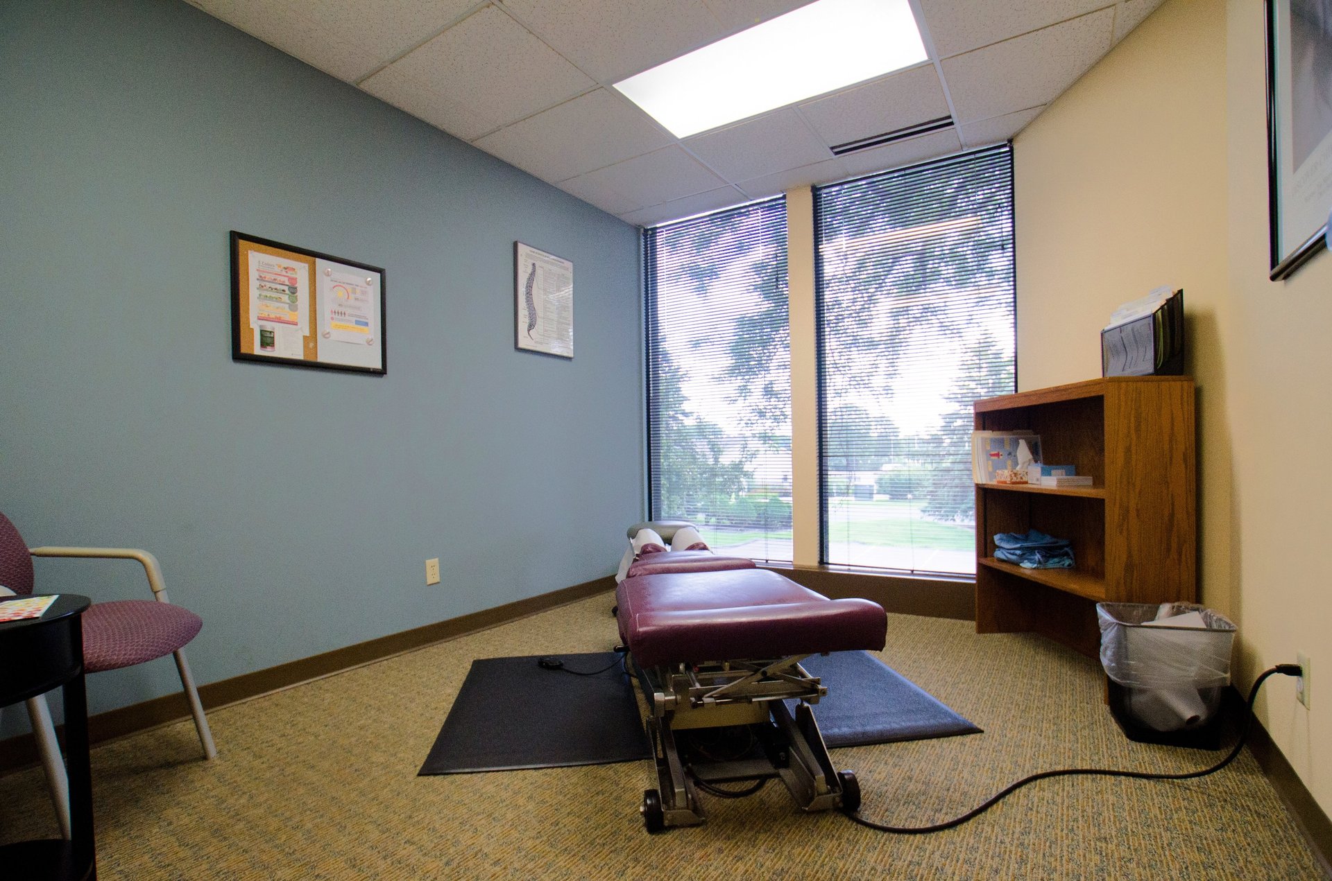 Clean chiropractic exam room with a treatment table, a bookcase of supplies, and a single chair.