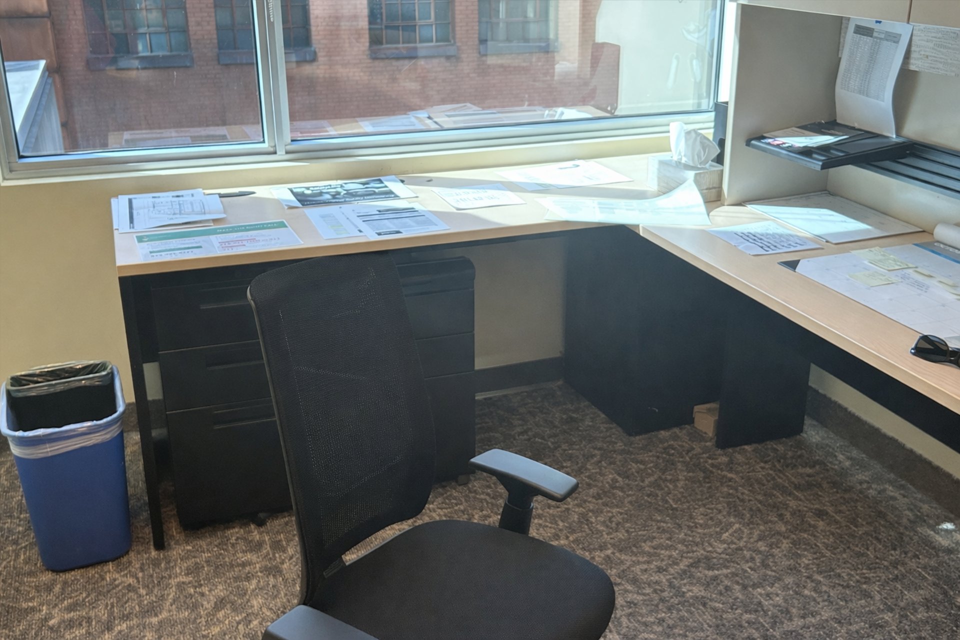 Office workspace with desk, chair, paperwork, and large window overlooking brick building