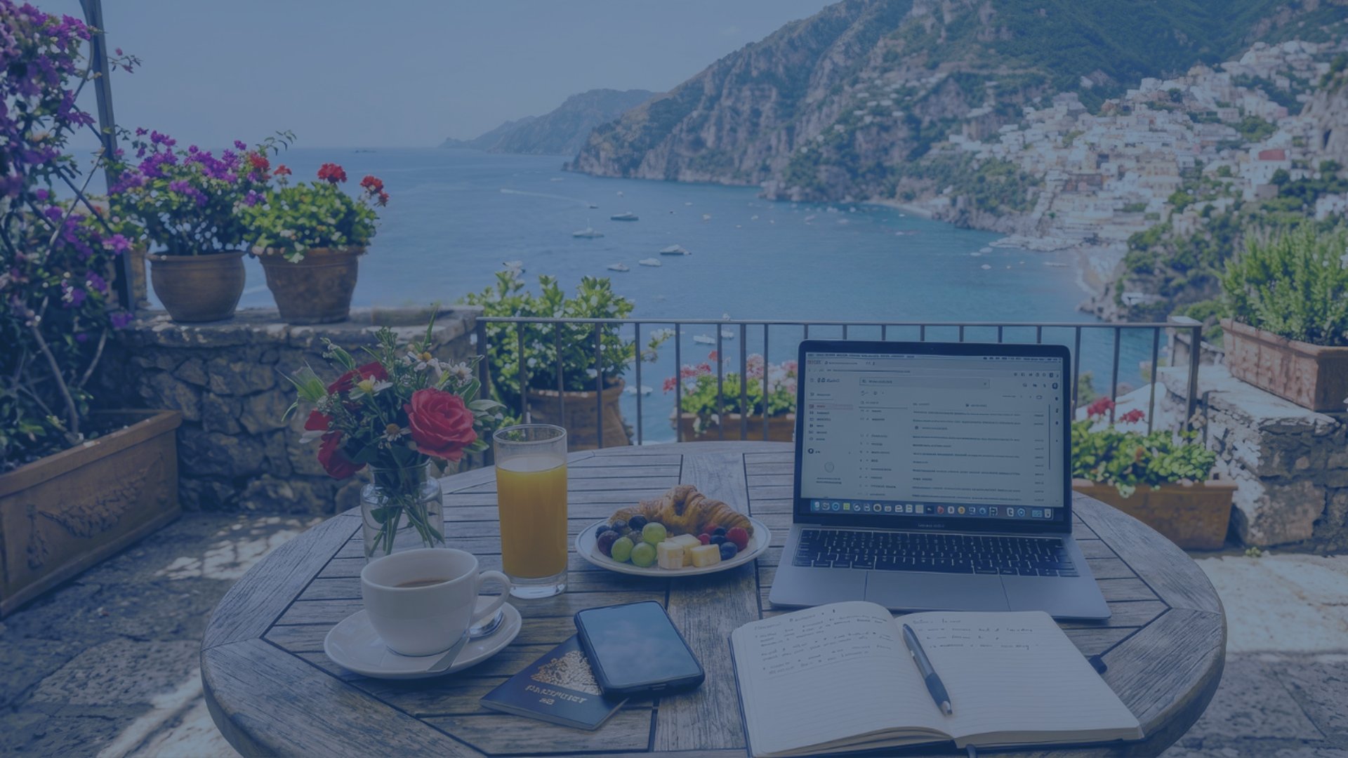 a work-travel lifestyle scene on a sunny Mediterranean terrace overlooking a curved coastal bay and hillside village. 