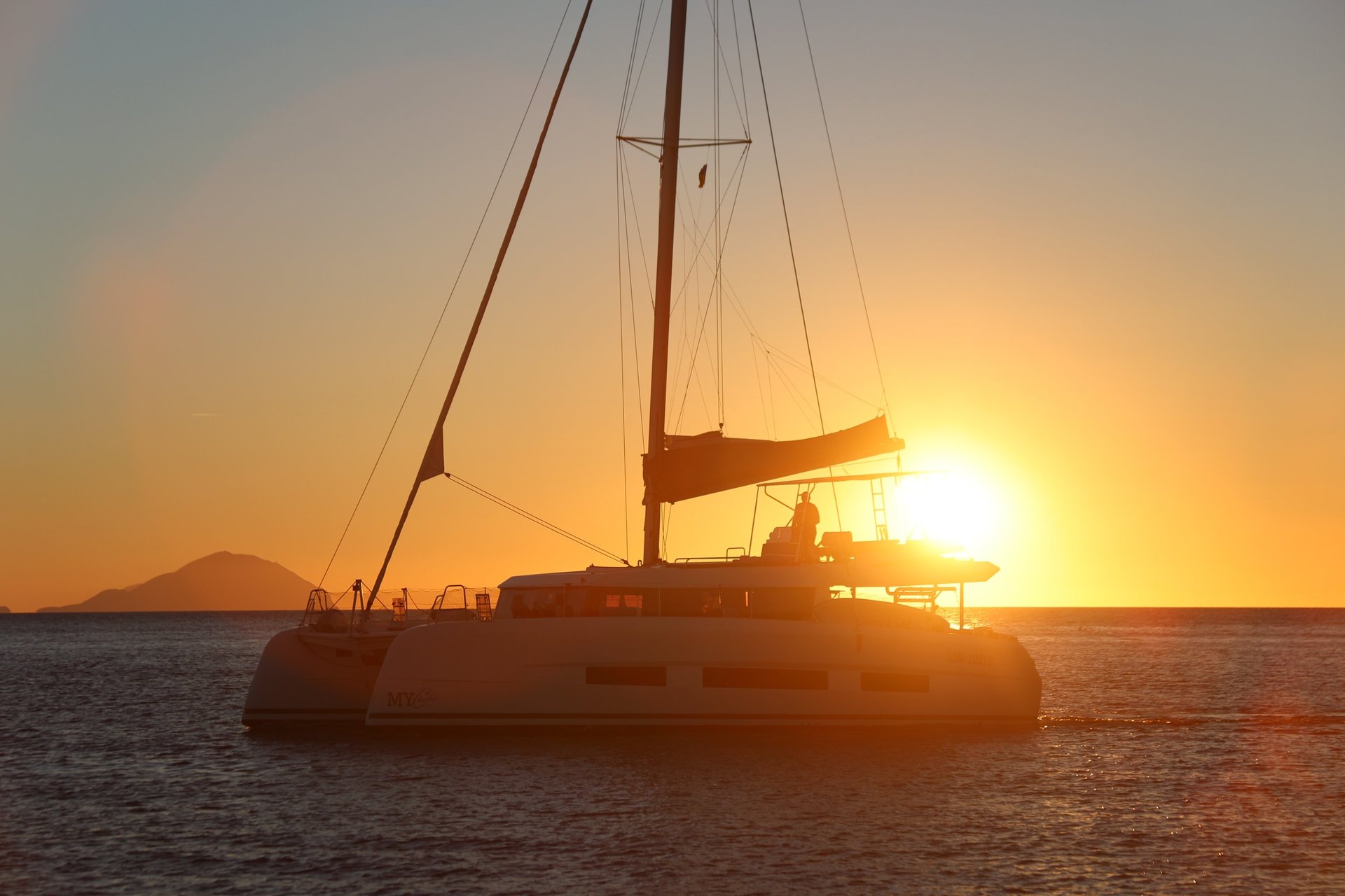 Sailboats glide across the calm ocean at sunset.